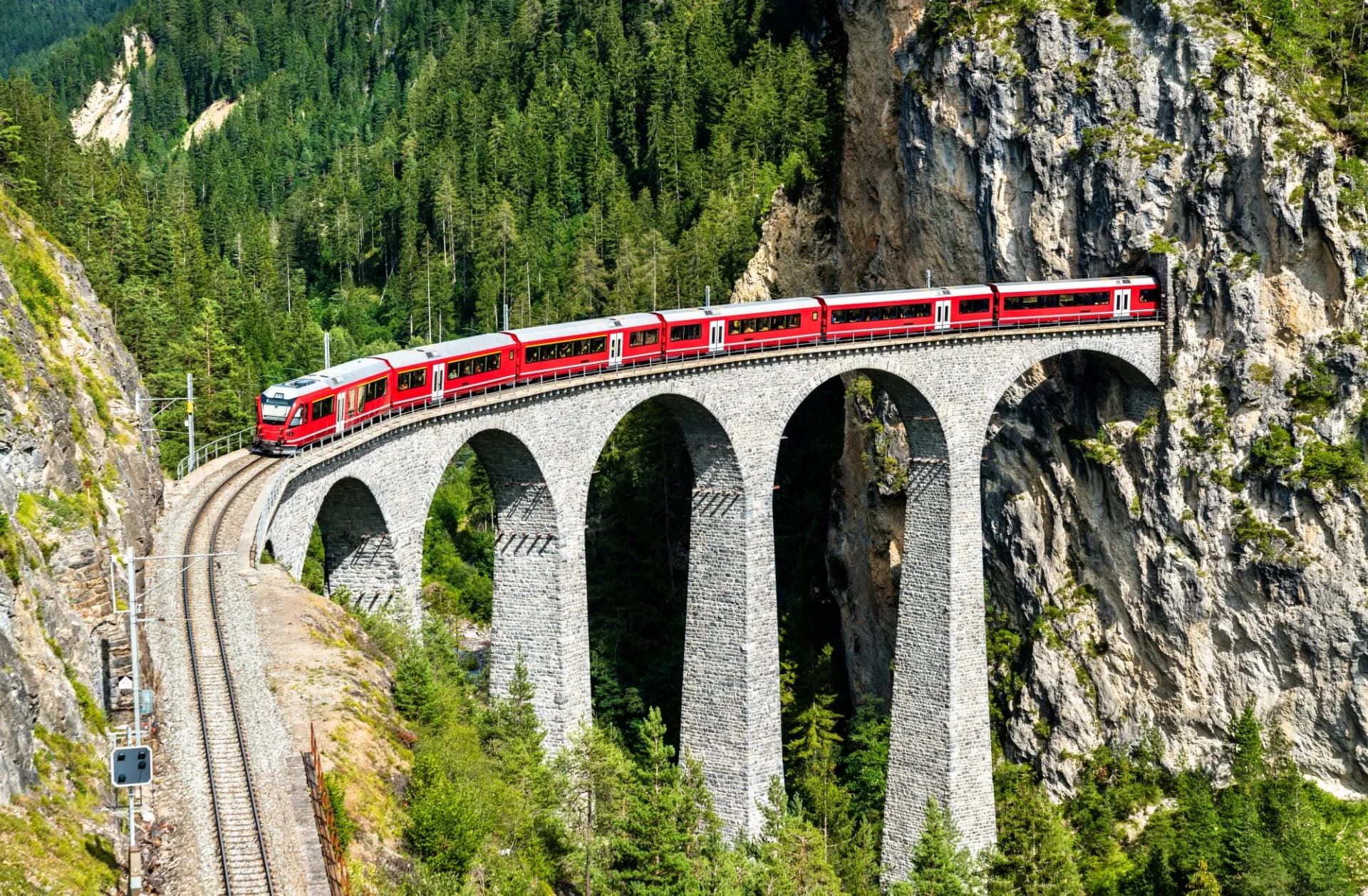 Red train crossing stone arched viaduct through lush green mountainside in summer