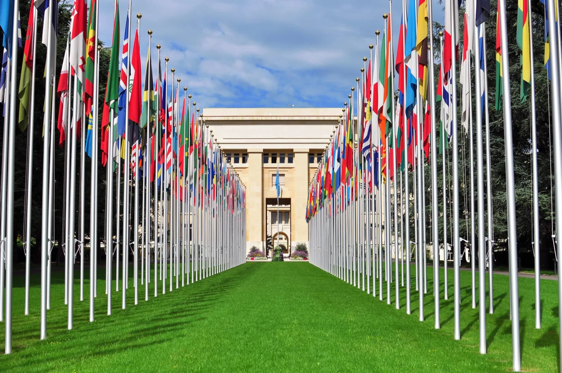 Rows of international flags flanking a grassy lawn leading to a large building in Geneva.