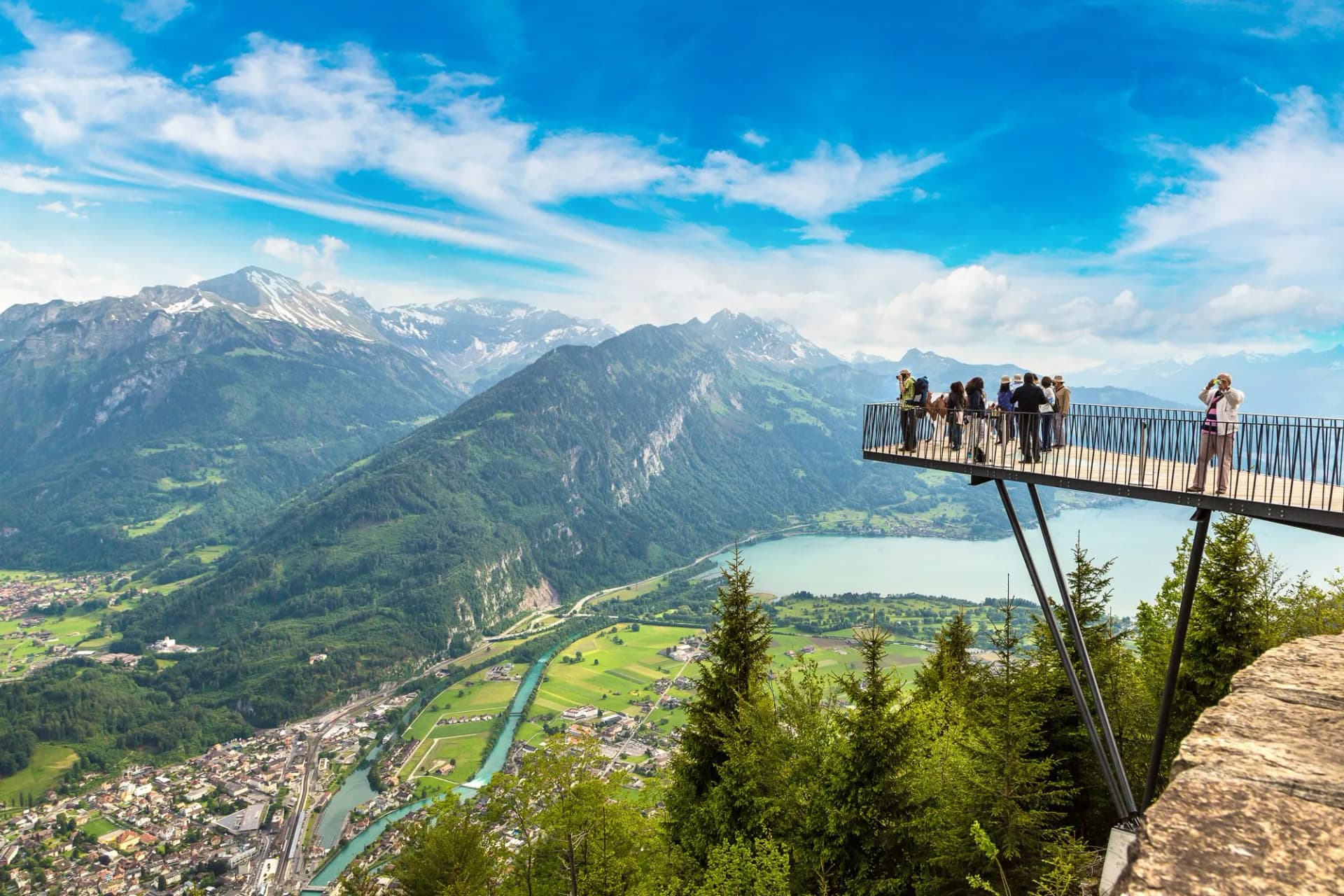 Observation deck visitors overlooking Interlaken town, lake, and green mountains under blue sky.