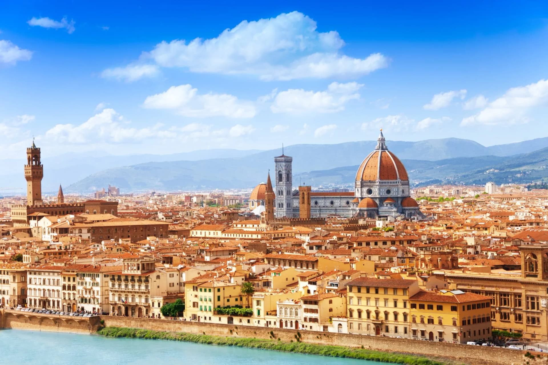 Cityscape of Florence with Duomo dome, Palazzo Vecchio tower, and river under blue sky.