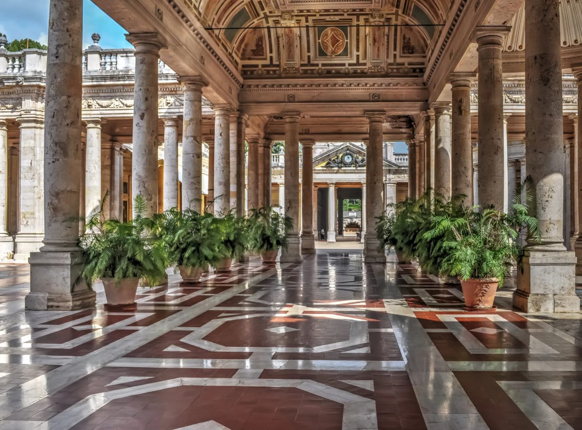 Colonnaded portico with ornate ceiling, patterned marble floor, and potted ferns in Montecatini Terme.