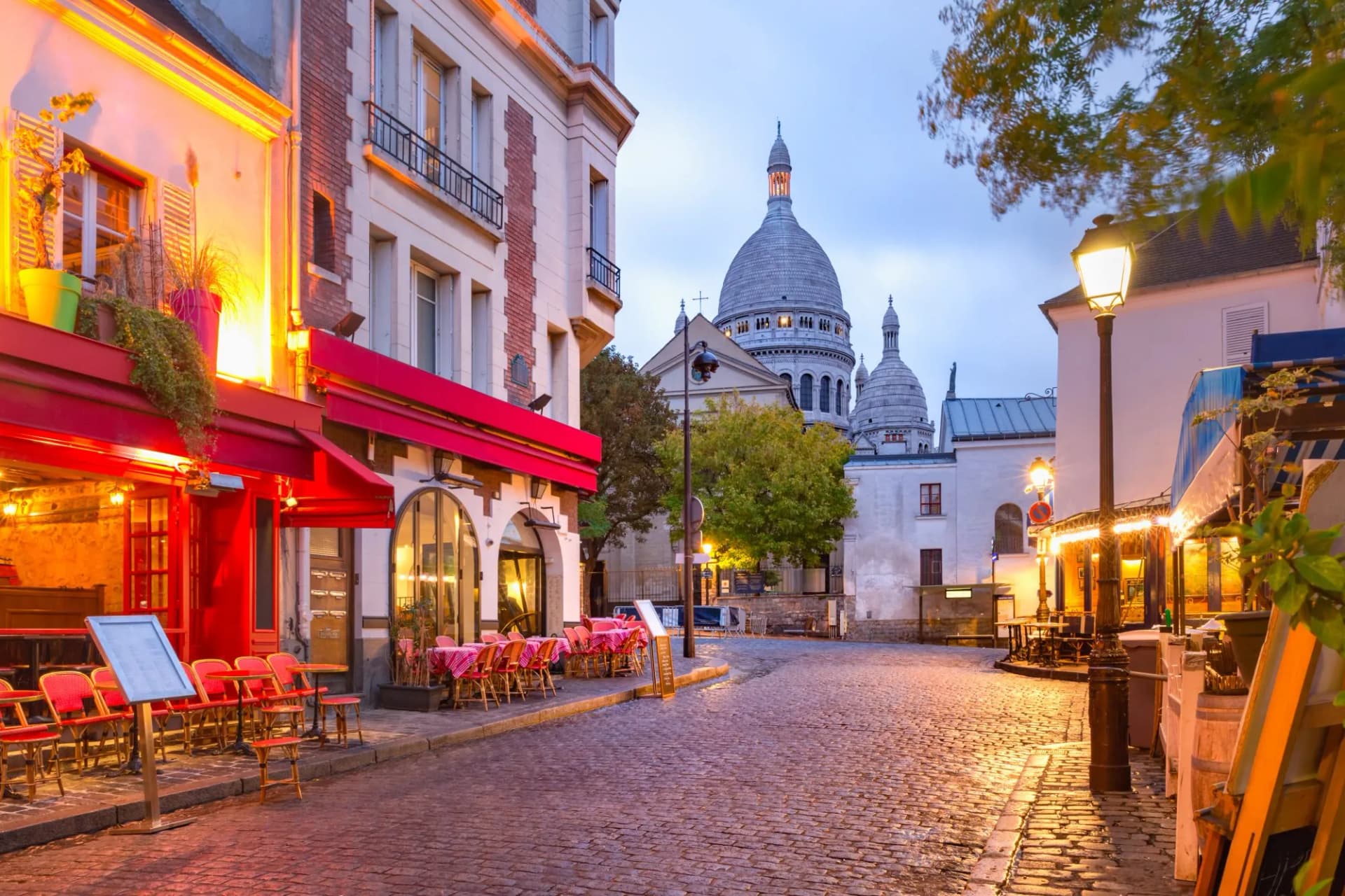 Cobblestone street leading to Sacré-Cœur Basilica in Montmartre, Paris at dusk with illuminated cafe.