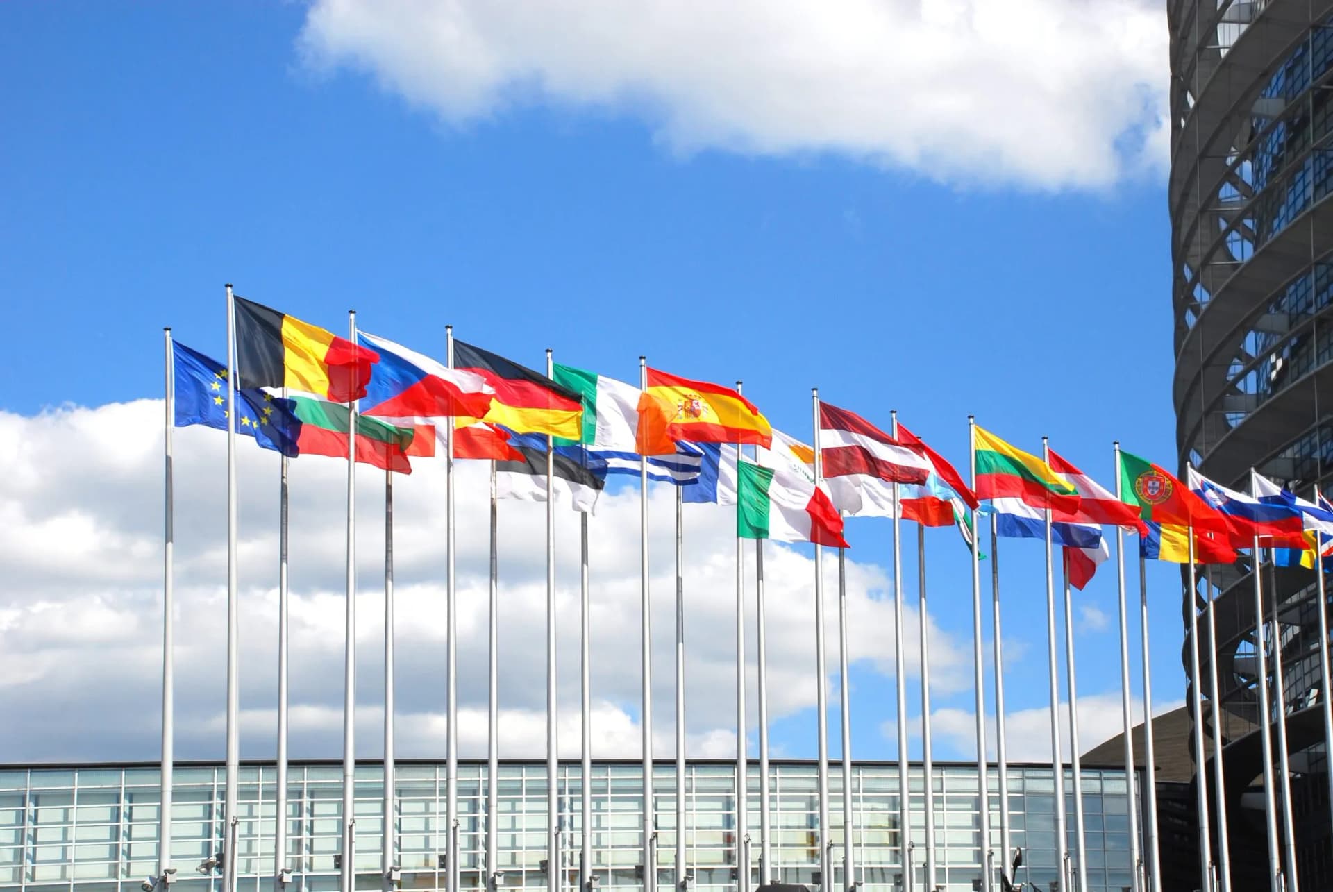 European Union and member state flags flying outside a modern building under a blue, cloudy sky in Strasbourg.
