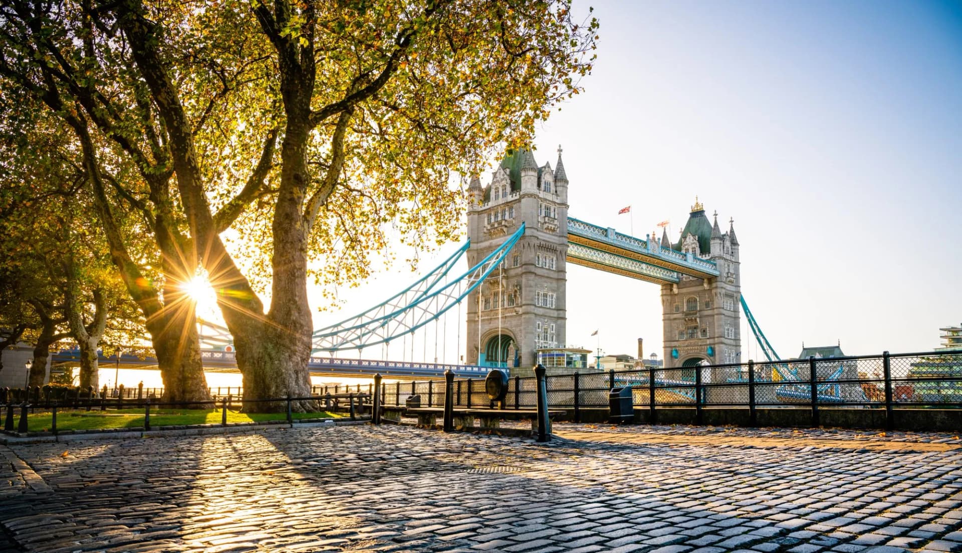Tower Bridge at sunrise in autumn with sun flare through trees over cobblestone path.