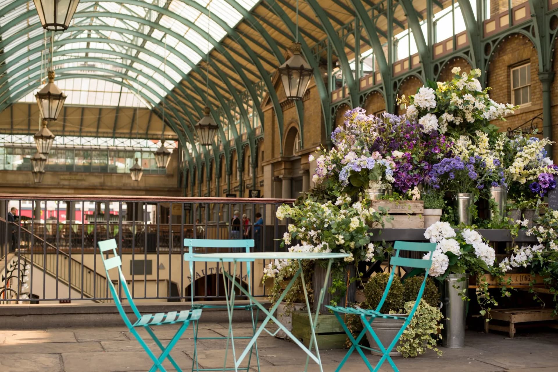 Outdoor cafe setting with teal chairs, flowers, and arched glass roof at Covent Garden Market.