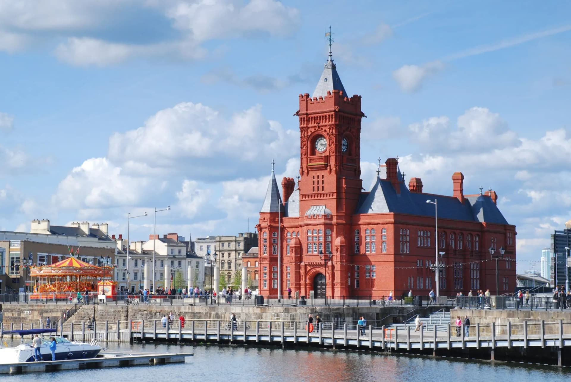 Red brick Pier Head Building in Cardiff Bay with a boat and carousel under blue sky.