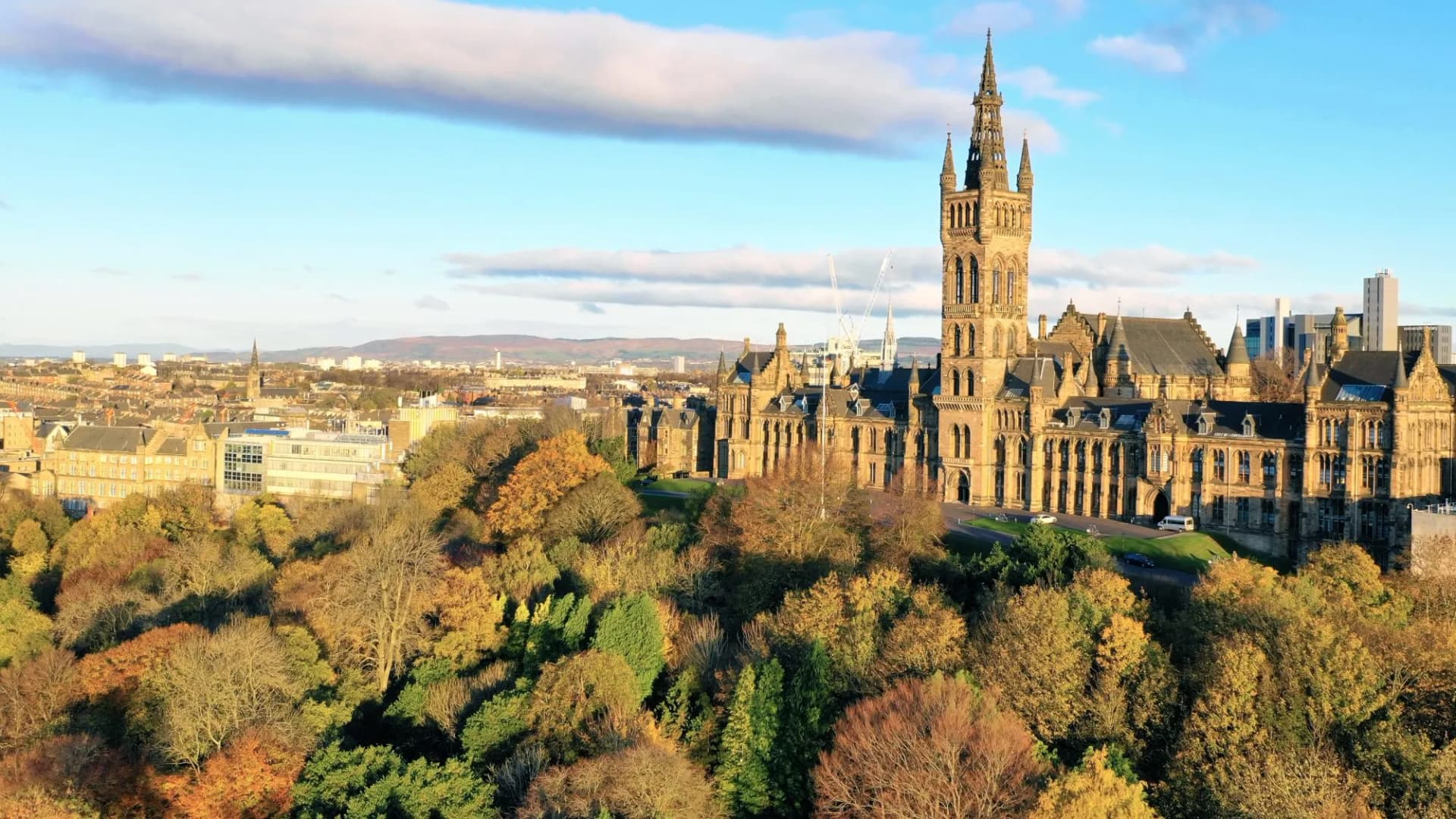 University of Glasgow main building with autumn trees and city skyline under blue sky.