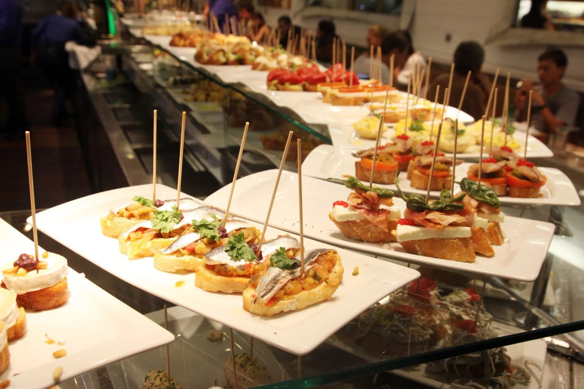 Assorted tapas and pintxos on display in a dimly lit bar with patrons in the background