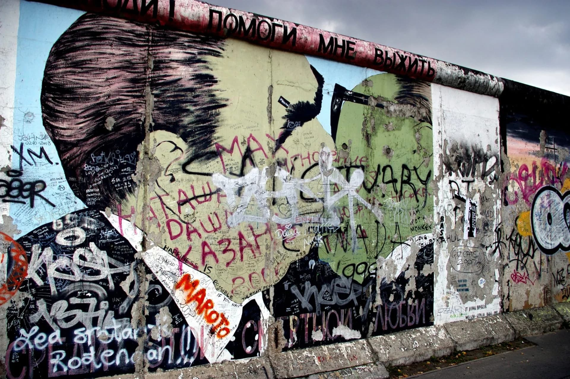 Graffiti mural on the Berlin Wall featuring a large painted portrait and Russian text under a gray sky.