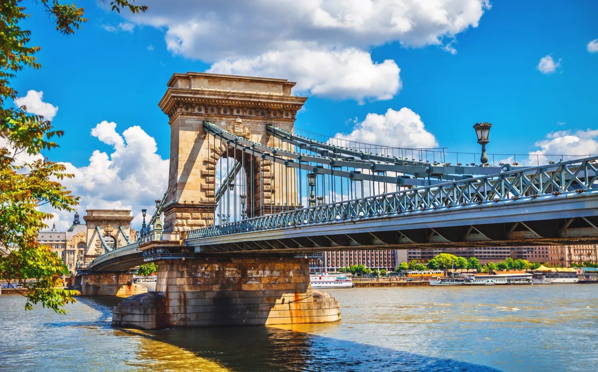 Chain Bridge in Budapest crossing the Danube River under a bright blue, cloudy sky.