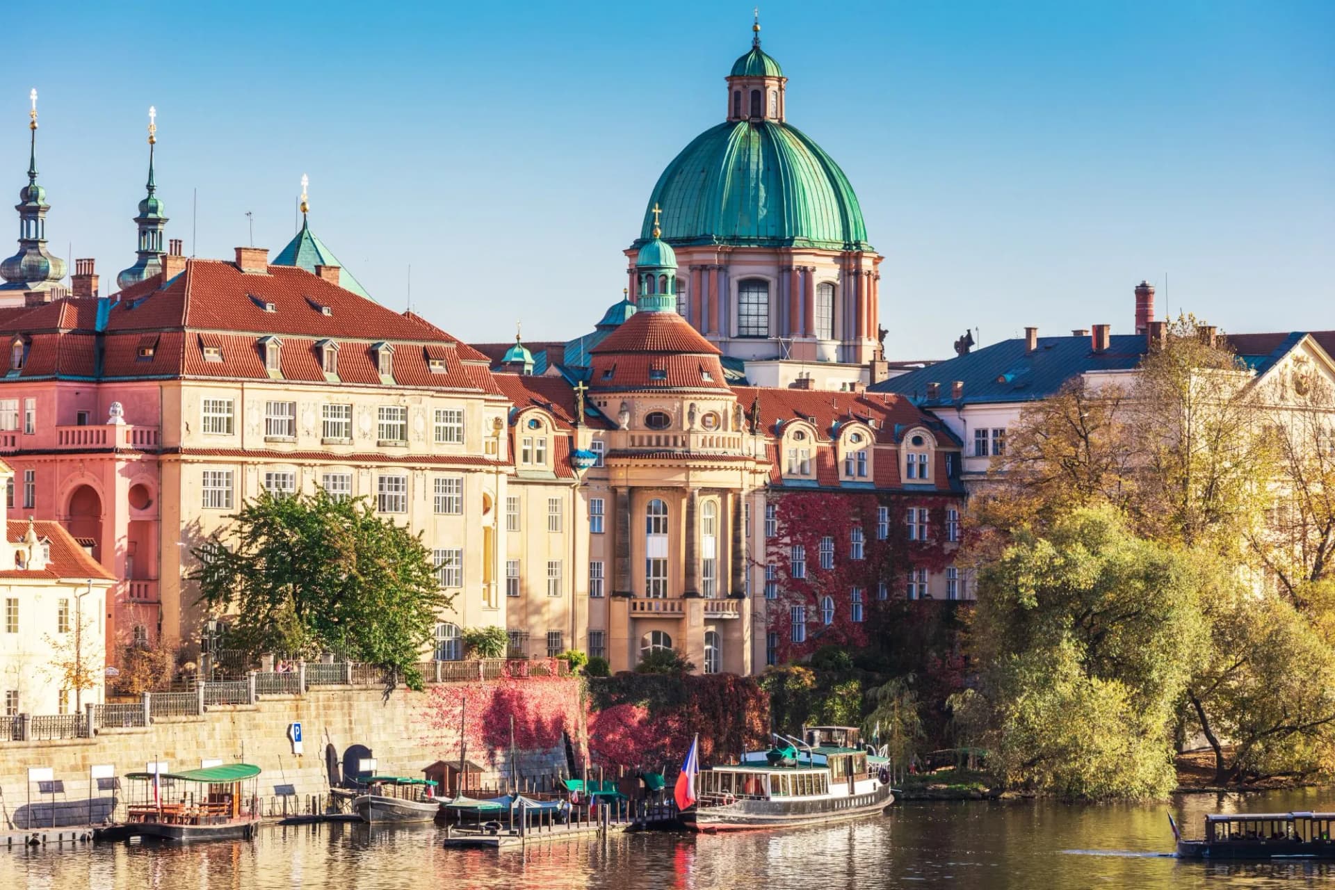 Old Prague buildings with red roofs and green dome along the Vltava River in autumn.