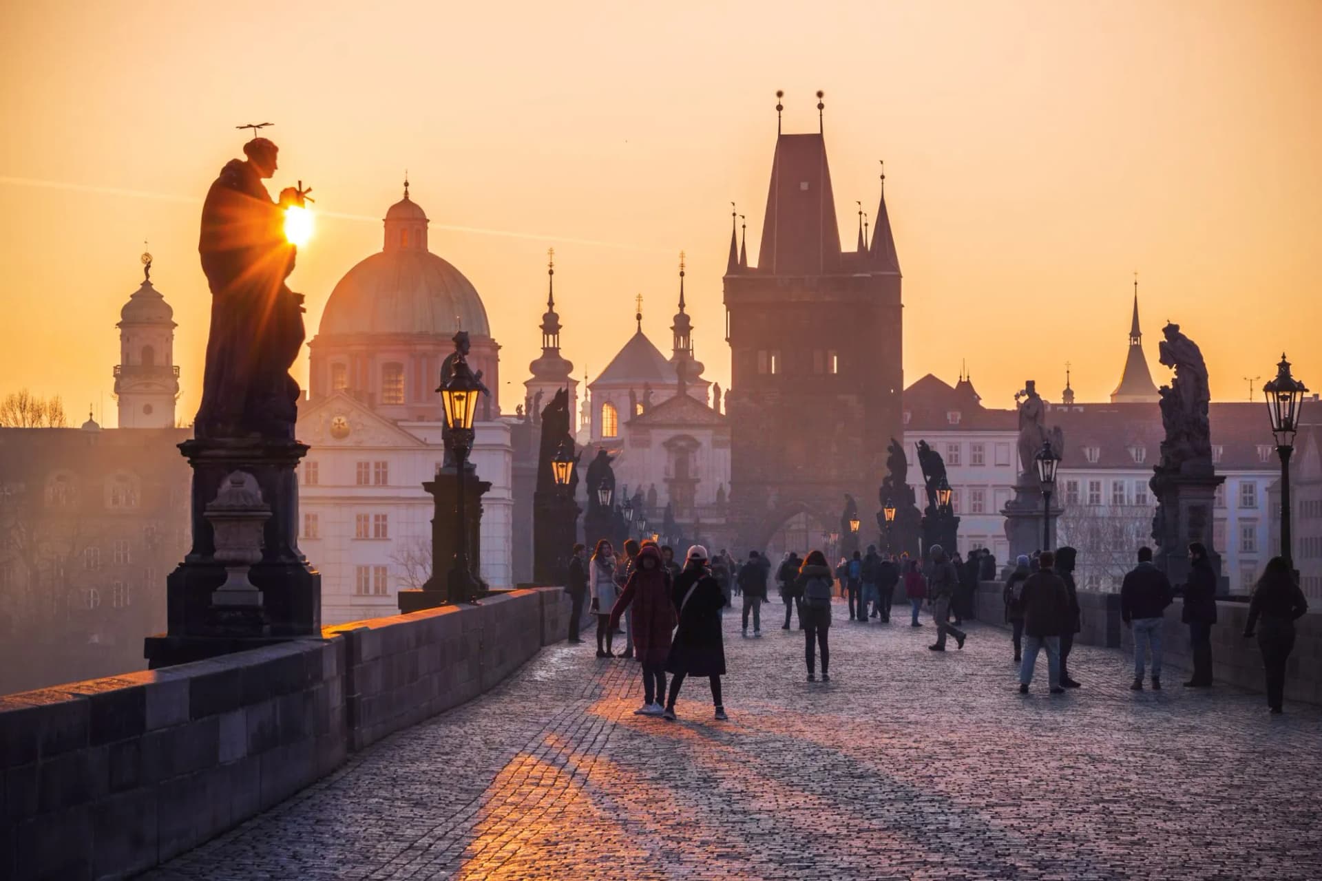 Statues and pedestrians on Charles Bridge in Prague at sunrise with city skyline.