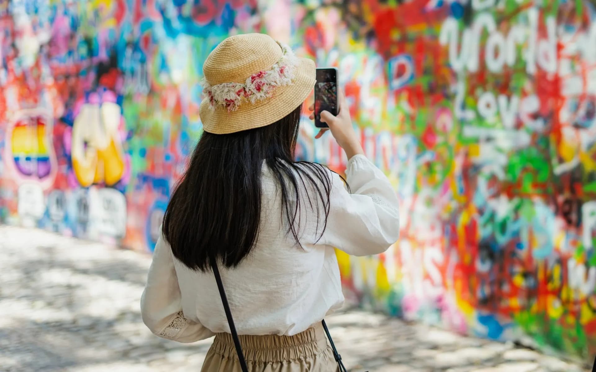 Tourist photographing colorful graffiti wall at Prague Lennon Wall with straw hat.