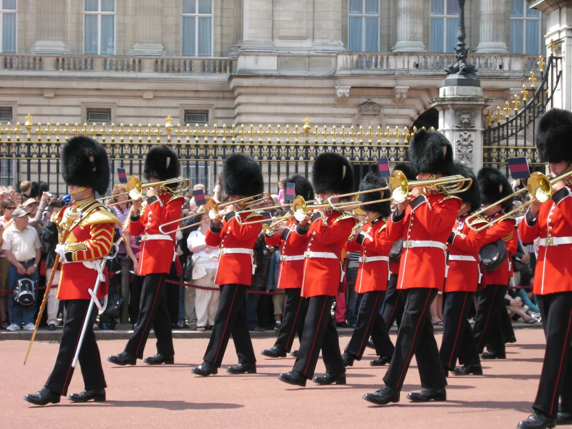 Changing of the Guard musicians in red tunics and bearskin hats marching past a palace gate.