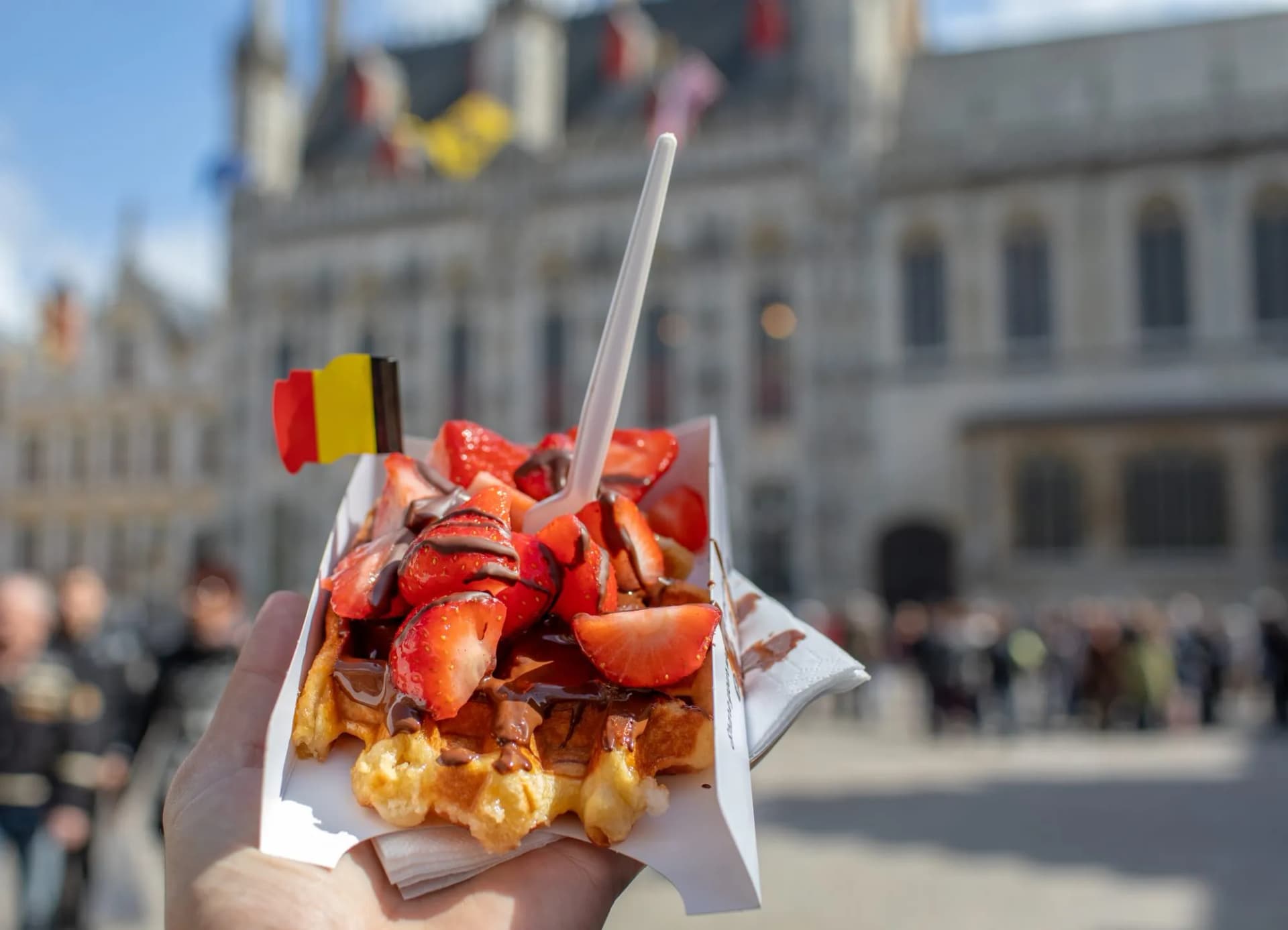 Brussels waffle with strawberries and chocolate, held outdoors near a historic building with a Belgian flag.