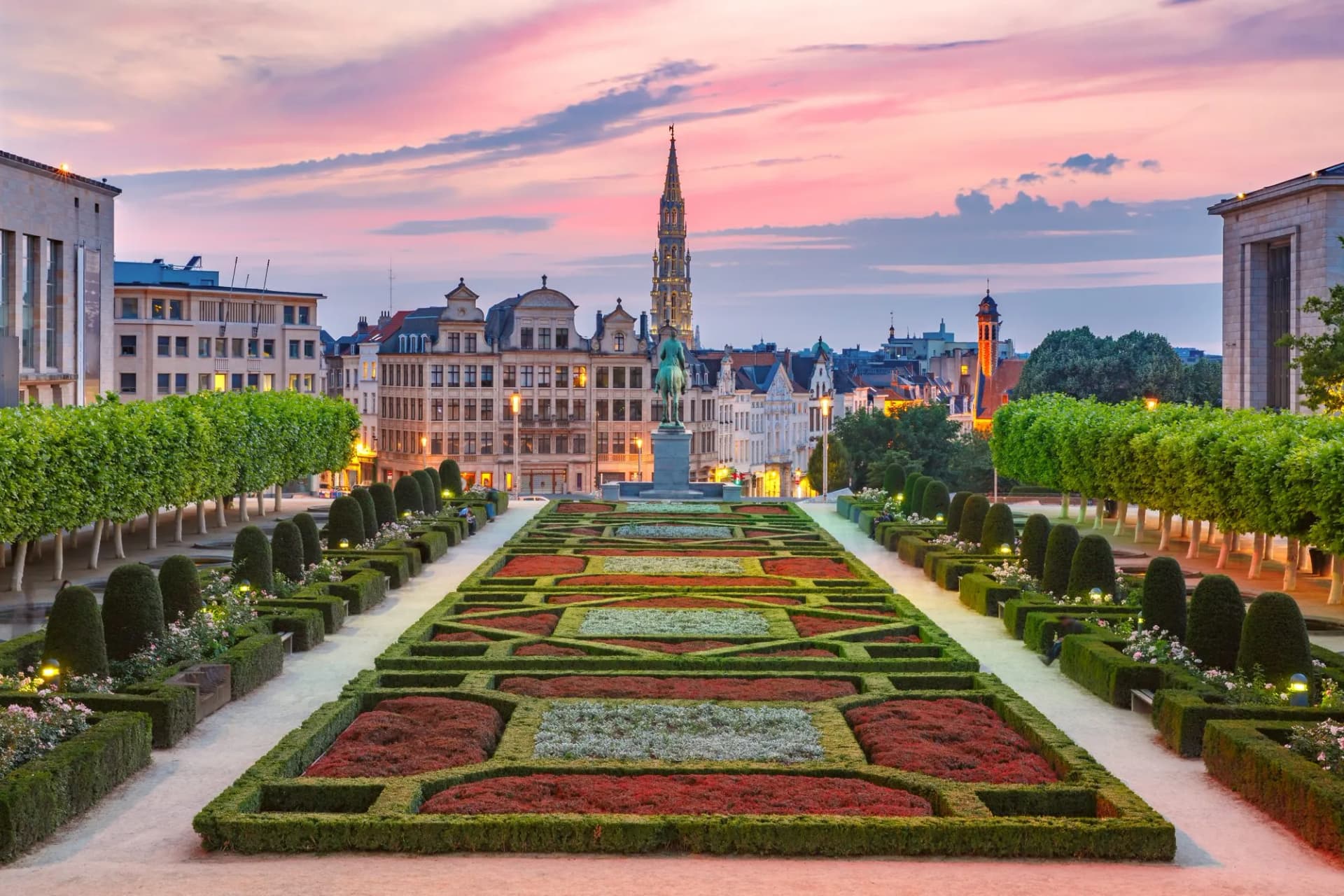 Formal gardens with patterned hedges leading to a city skyline and spire at sunset in Brussels.