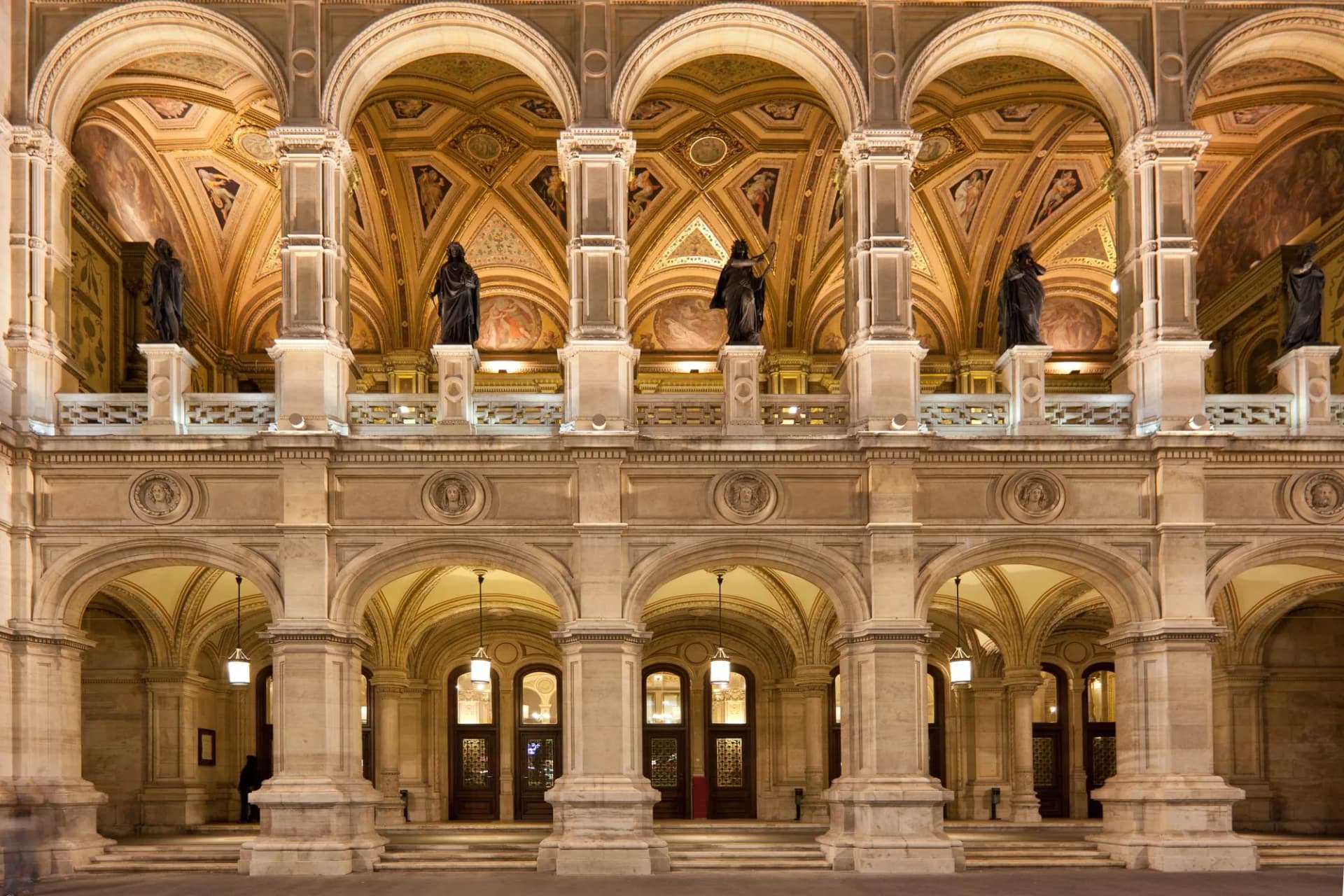 Illuminated arcade entrance of the Wiener Staatsoper at night with statues above.