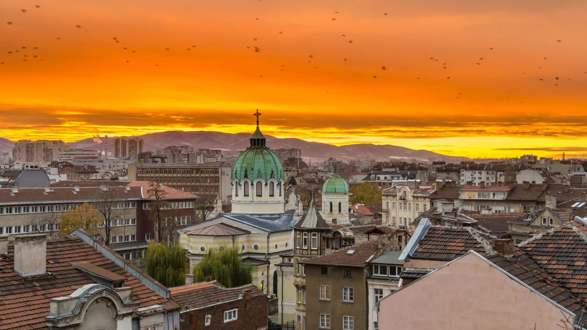 Aerial view of Sofia, Bulgaria cityscape with church domes against an intense orange sunset and mountains.