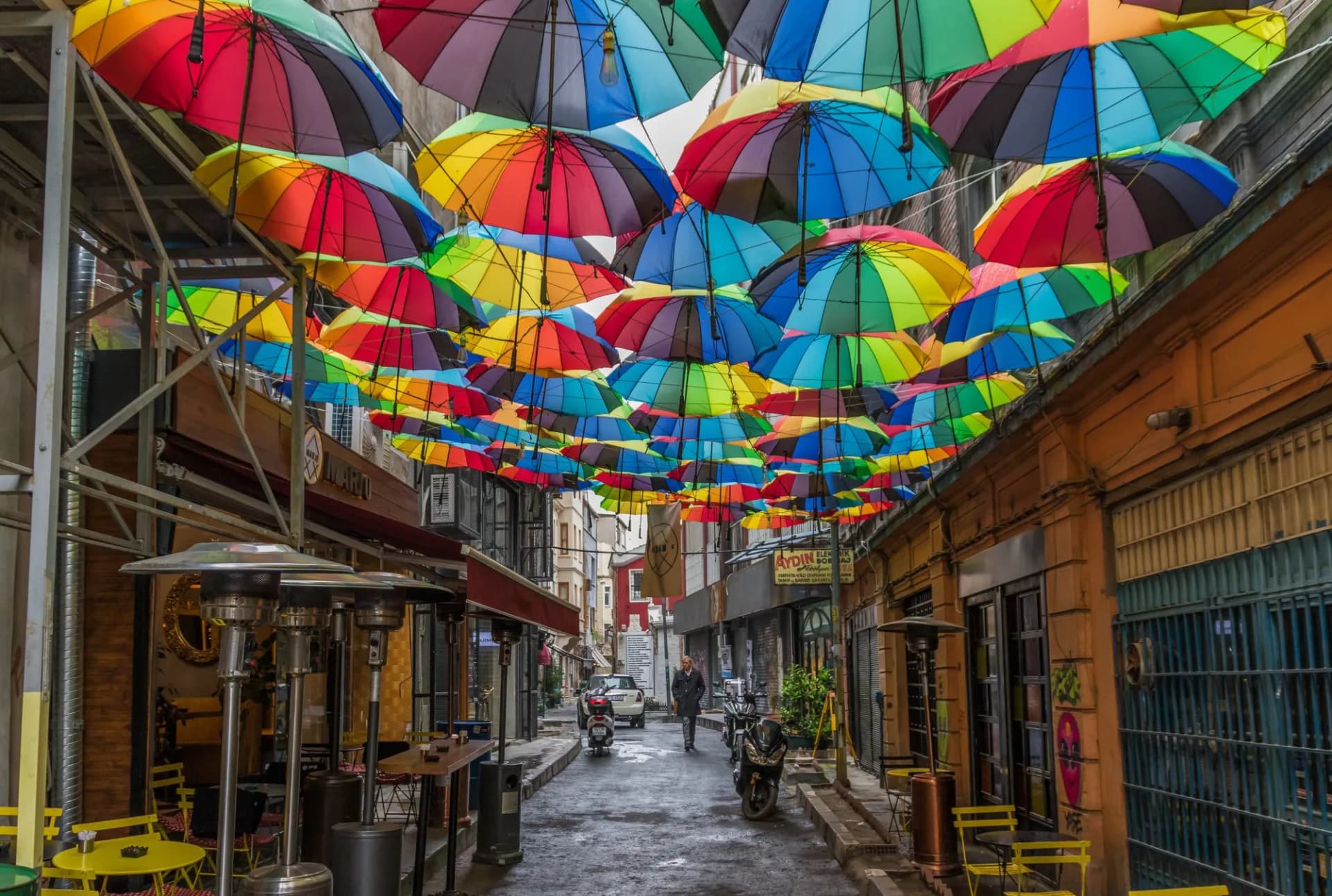 Colorful umbrellas suspended over a narrow street with outdoor cafe seating and heaters.