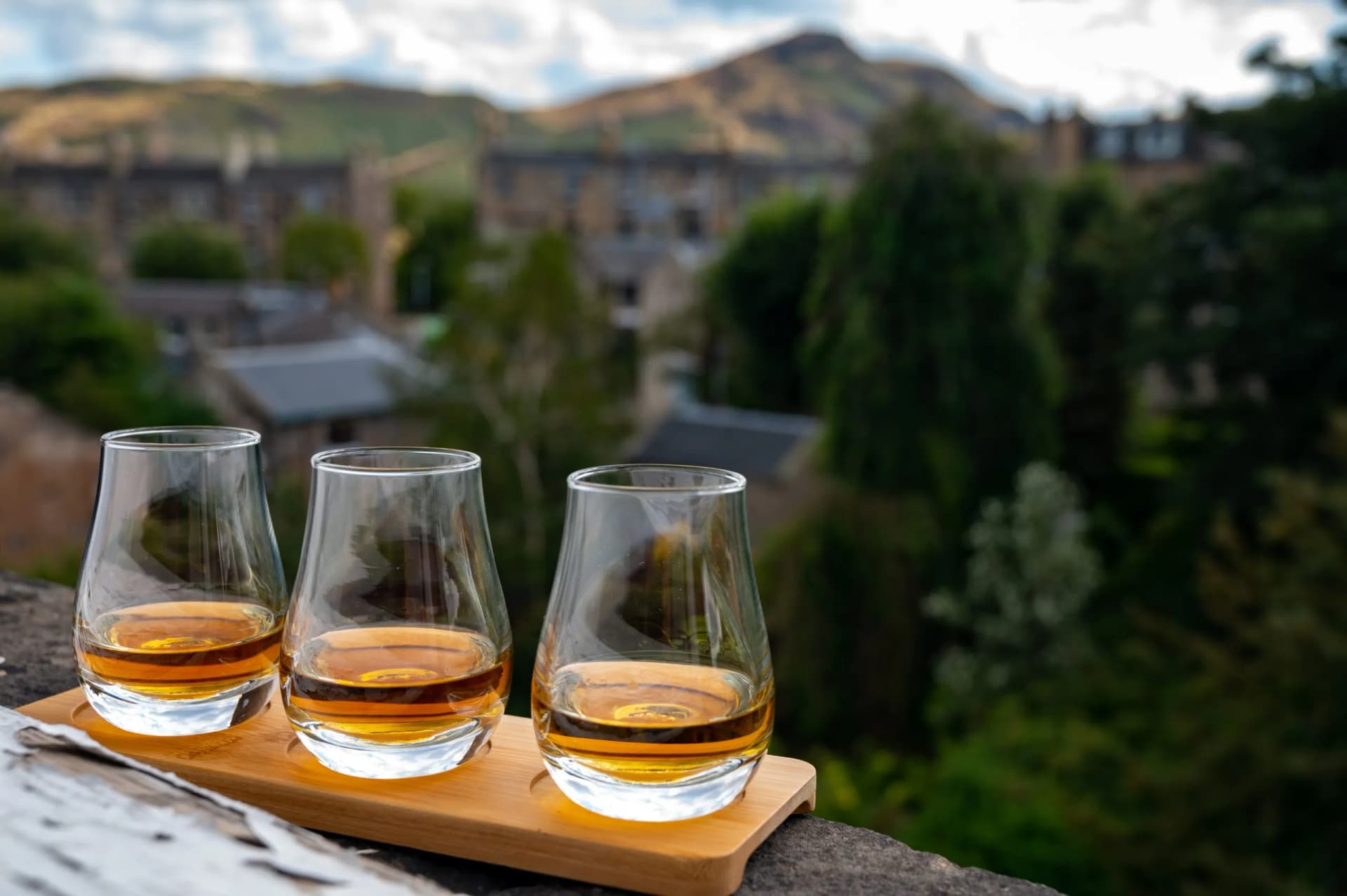 Three glasses of whisky on a wooden tray overlooking Edinburgh with Arthur's Seat in the background.