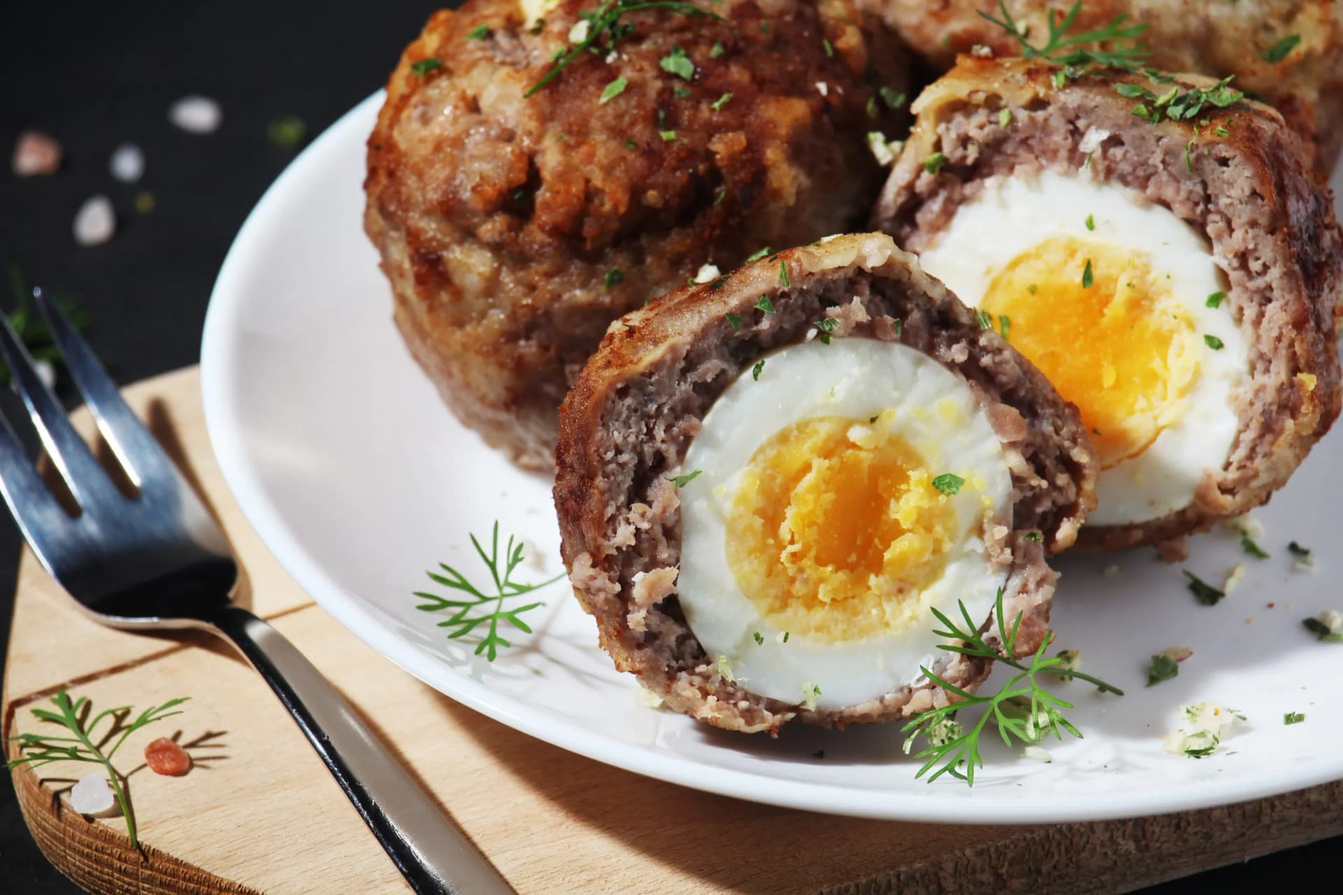 Scotch eggs sliced in half showing soft yolk, garnished with dill on a white plate with a fork.