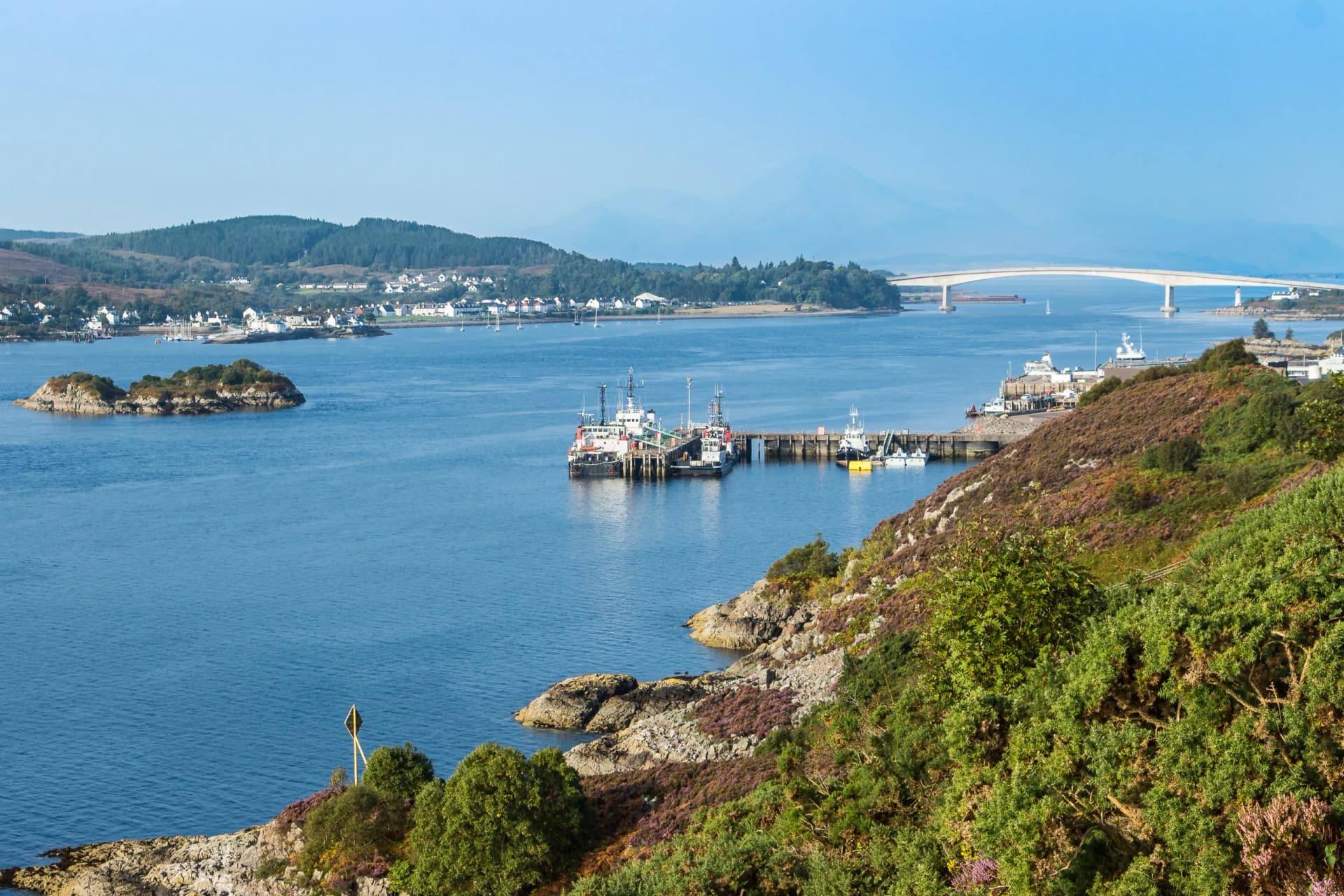 Coastal view with bridge, harbor, boats, and heather-covered hillside near Skye.