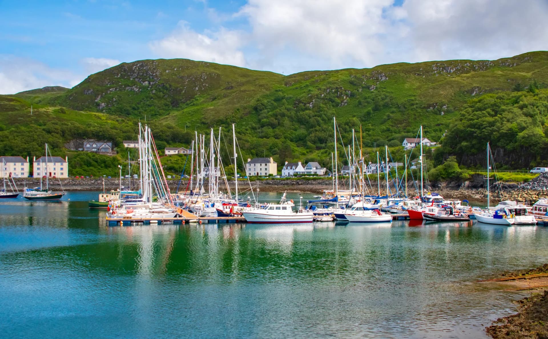 Sailboats and motorboats moored in Mallaig Harbour with green hills and white houses backdrop.