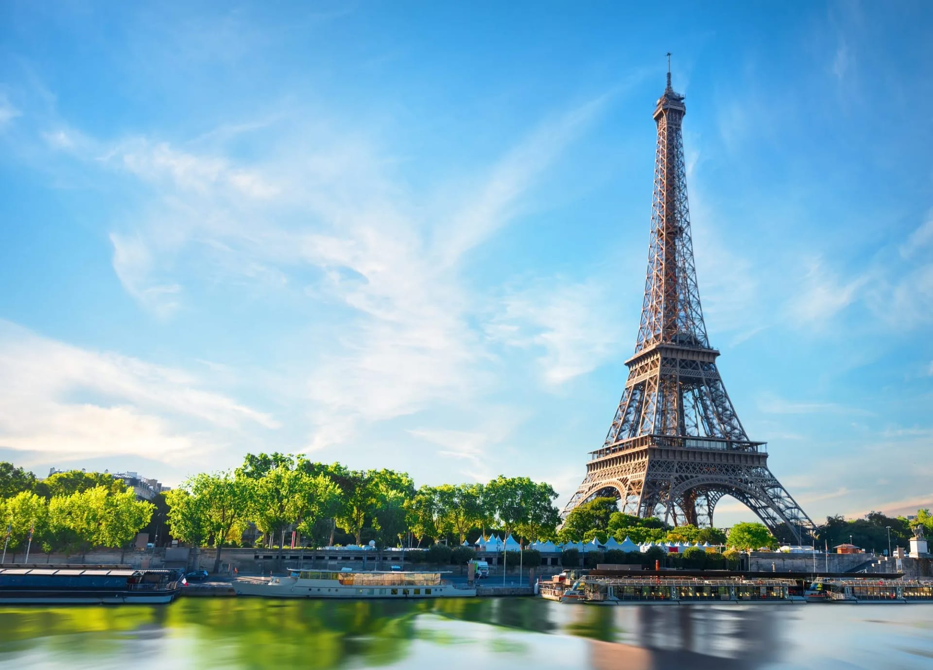 Eiffel Tower in Paris beside the Seine River with tour boats and green trees under blue sky.