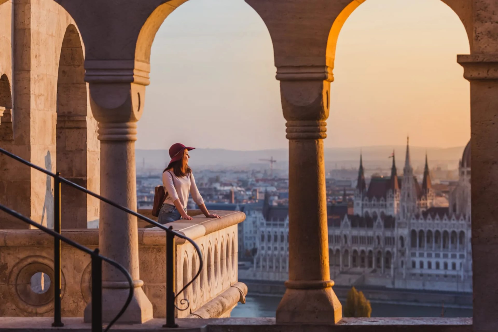 Woman with hat viewing Hungarian Parliament Building from Fisherman's Bastion arches at sunset.