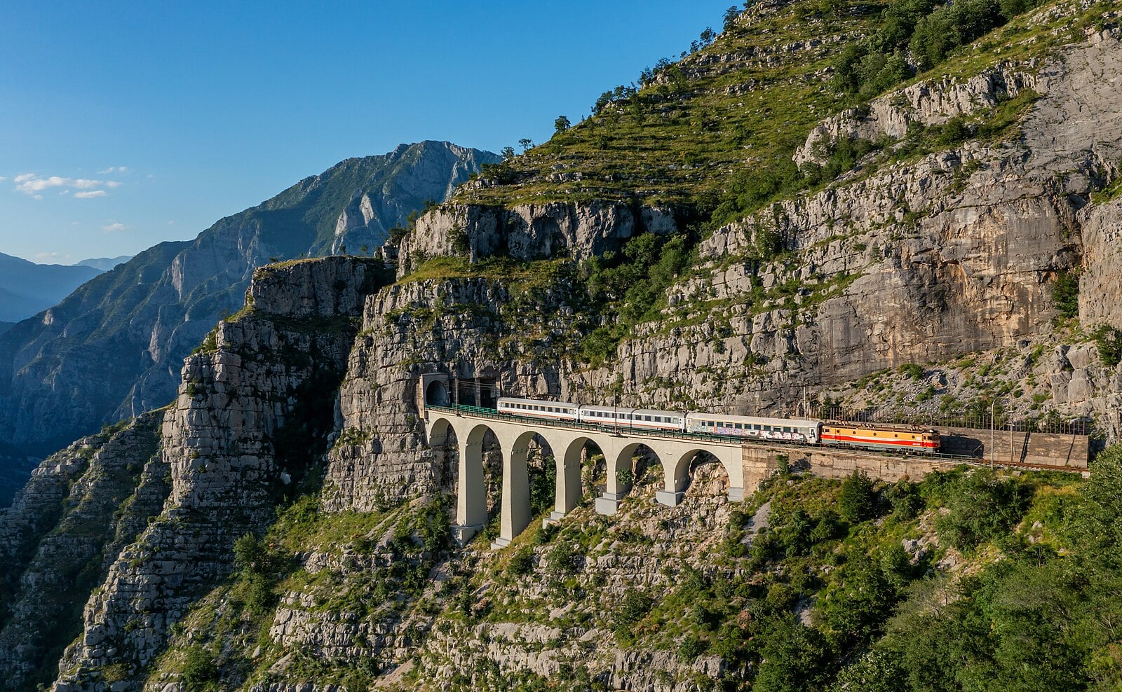 Train crossing arched viaduct built into steep, rocky mountainside on Belgrade-Bar line.