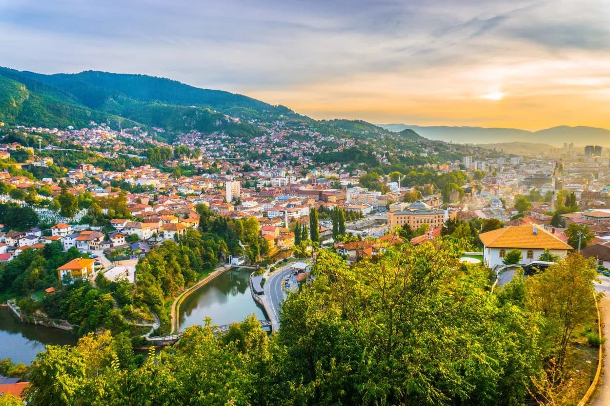 Panoramic view of Sarajevo city nestled in green mountains at sunset with a river bend.