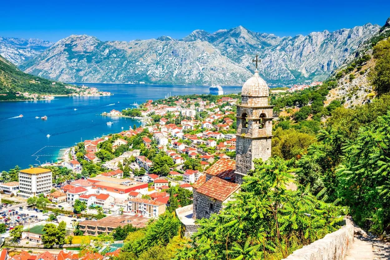 View from walls of Kotor showing stone bell tower, red-roofed town, and mountains surrounding the bay.