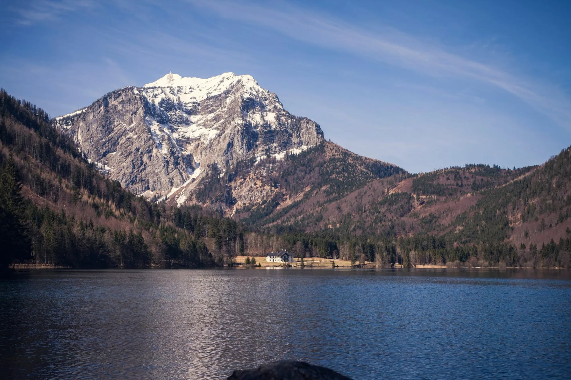 Alpine lake in Austria with snow-capped mountain and lakeside house under blue sky.