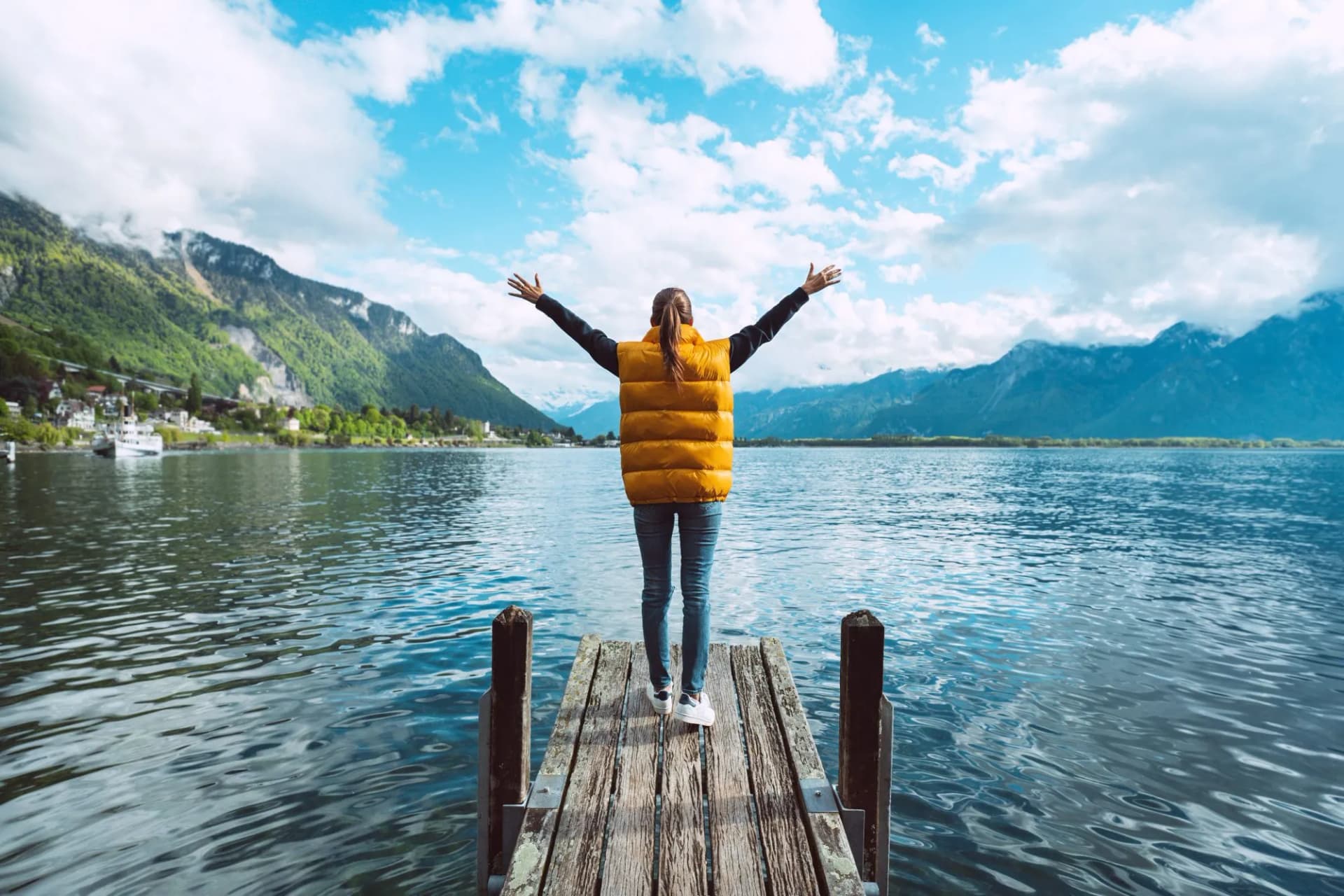 Woman with arms raised on wooden dock enjoying view of Geneva Lake and mountains in Switzerland.