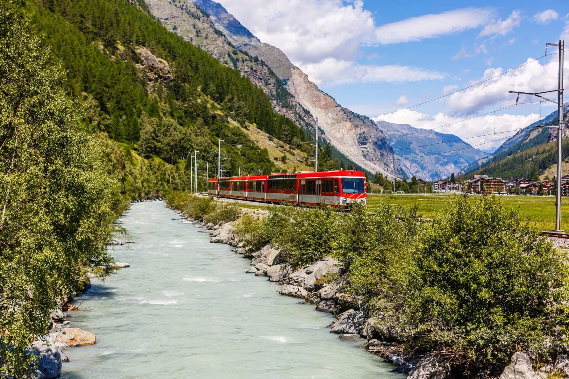 Red and white train crosses Alps railway beside a rushing river in the mountains.