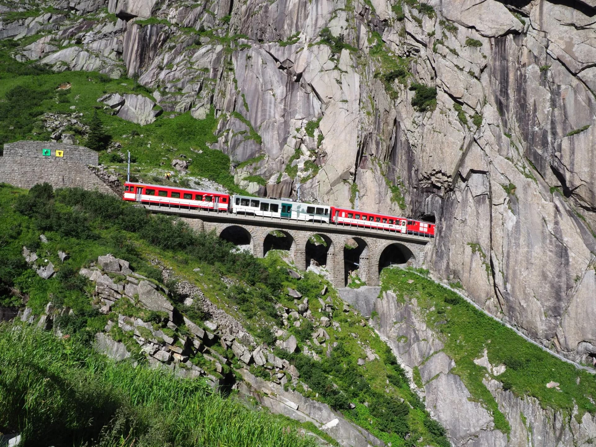 Red express train crossing a stone viaduct on the Gotthard Railway bridge in the Swiss Alps.