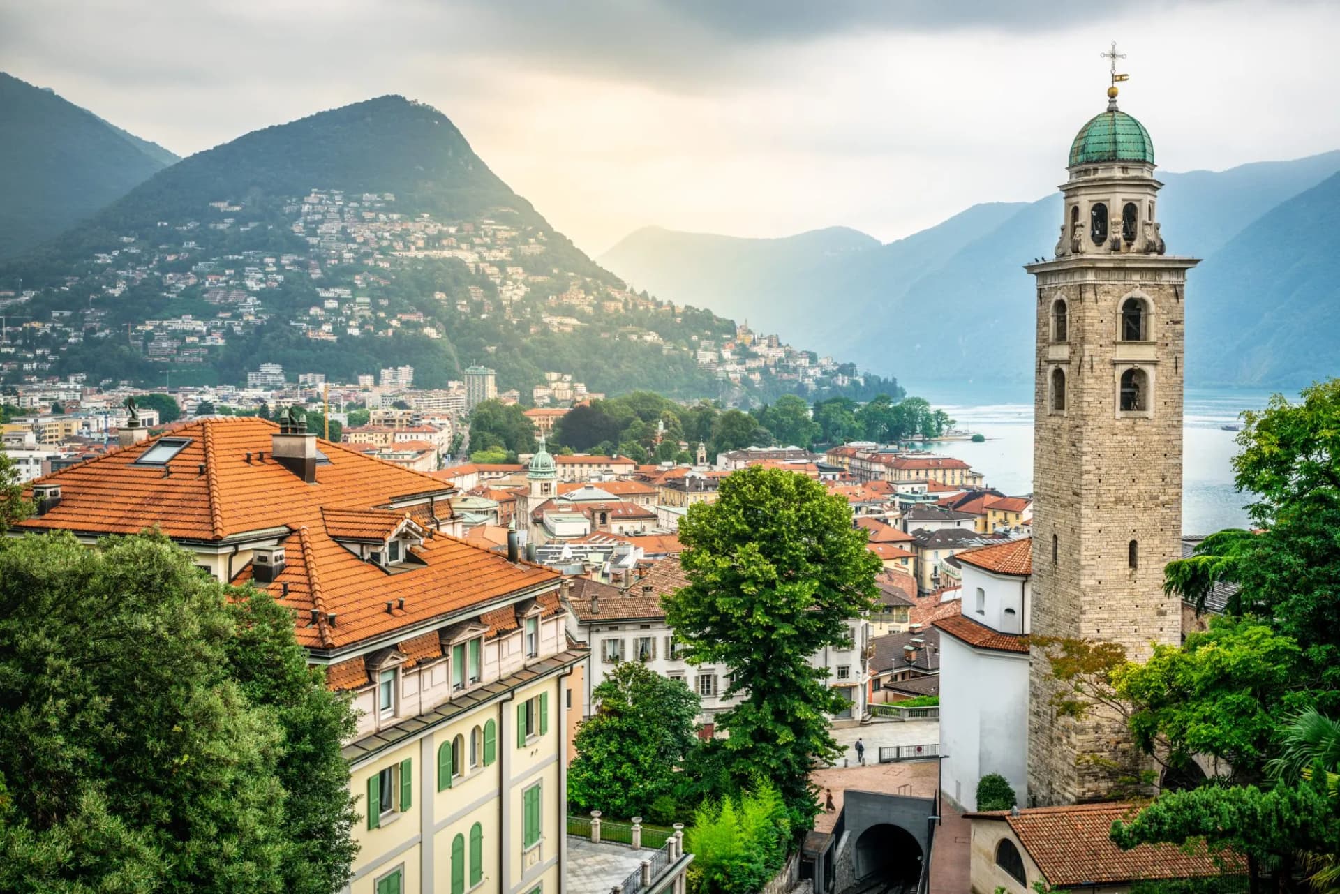 Cathedral of Saint Lawrence bell tower overlooking Lugano cityscape, lake, and mountains in Switzerland.