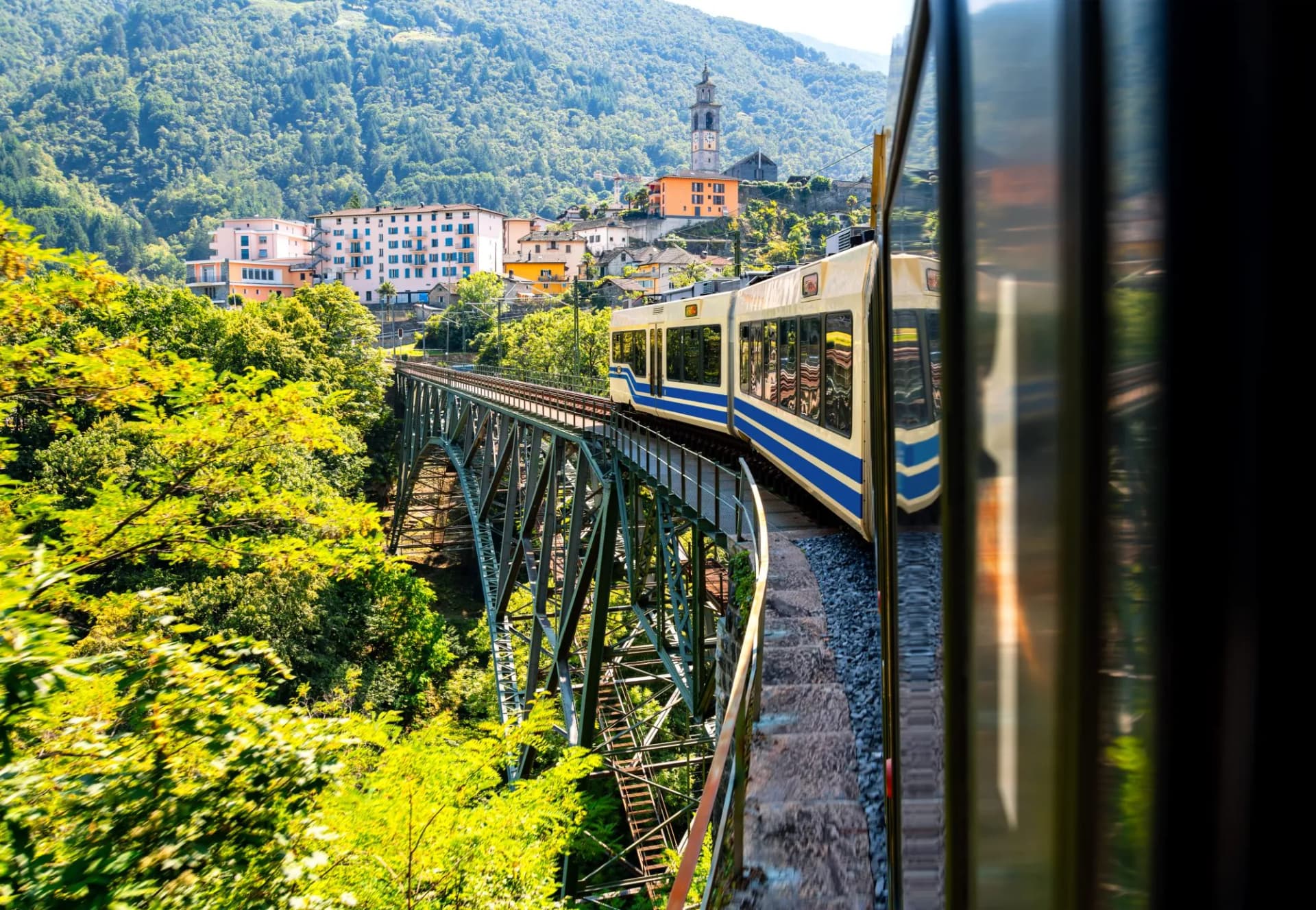 Centovalli Railway train crossing a steel bridge near Intragna with a village on a forested hillside.
