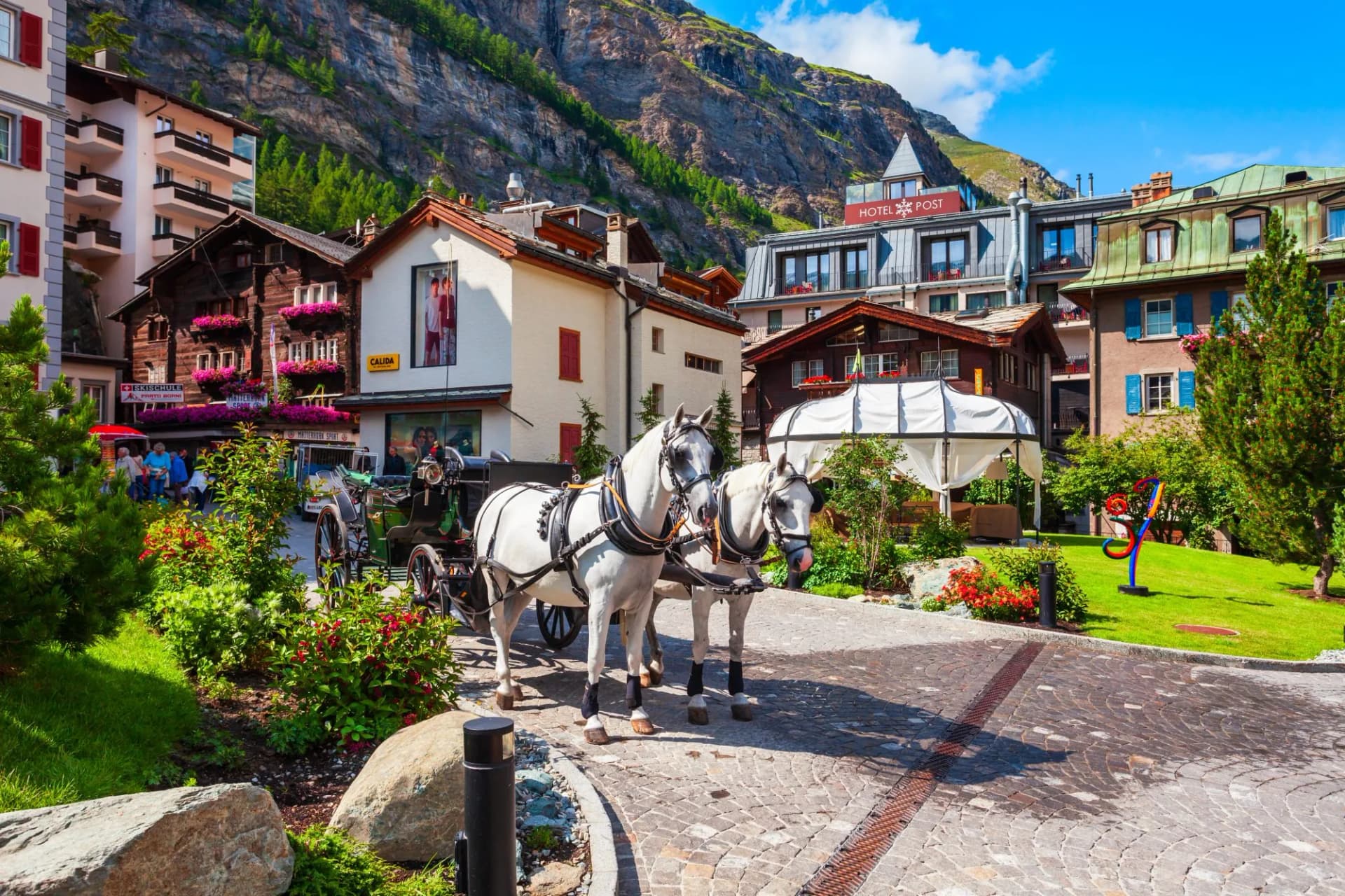 Two white horses pull a carriage past traditional houses beneath a steep mountain in Zermatt, Switzerland.