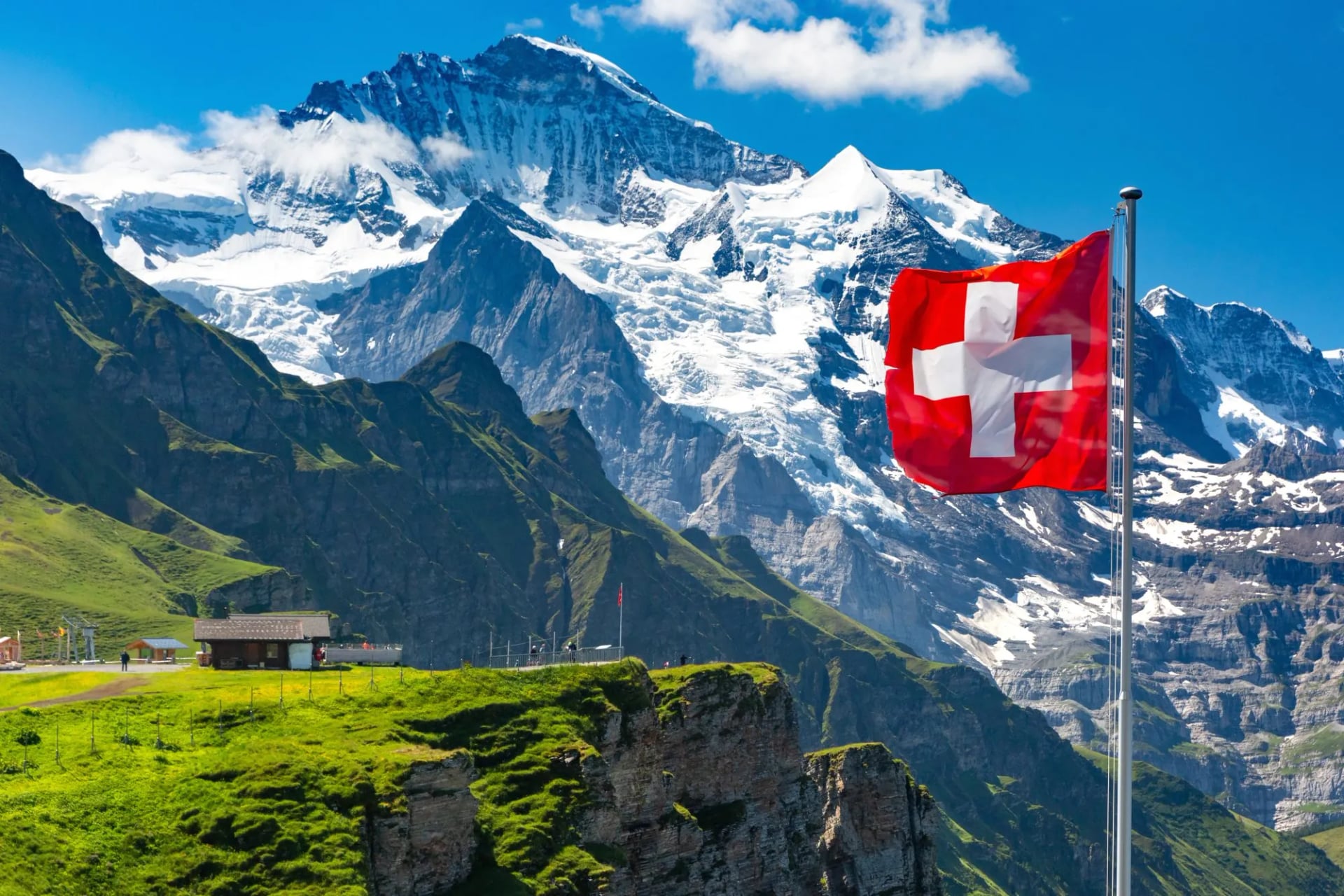 Swiss flag flying with snow-capped mountains and green alpine cliffs in Switzerland