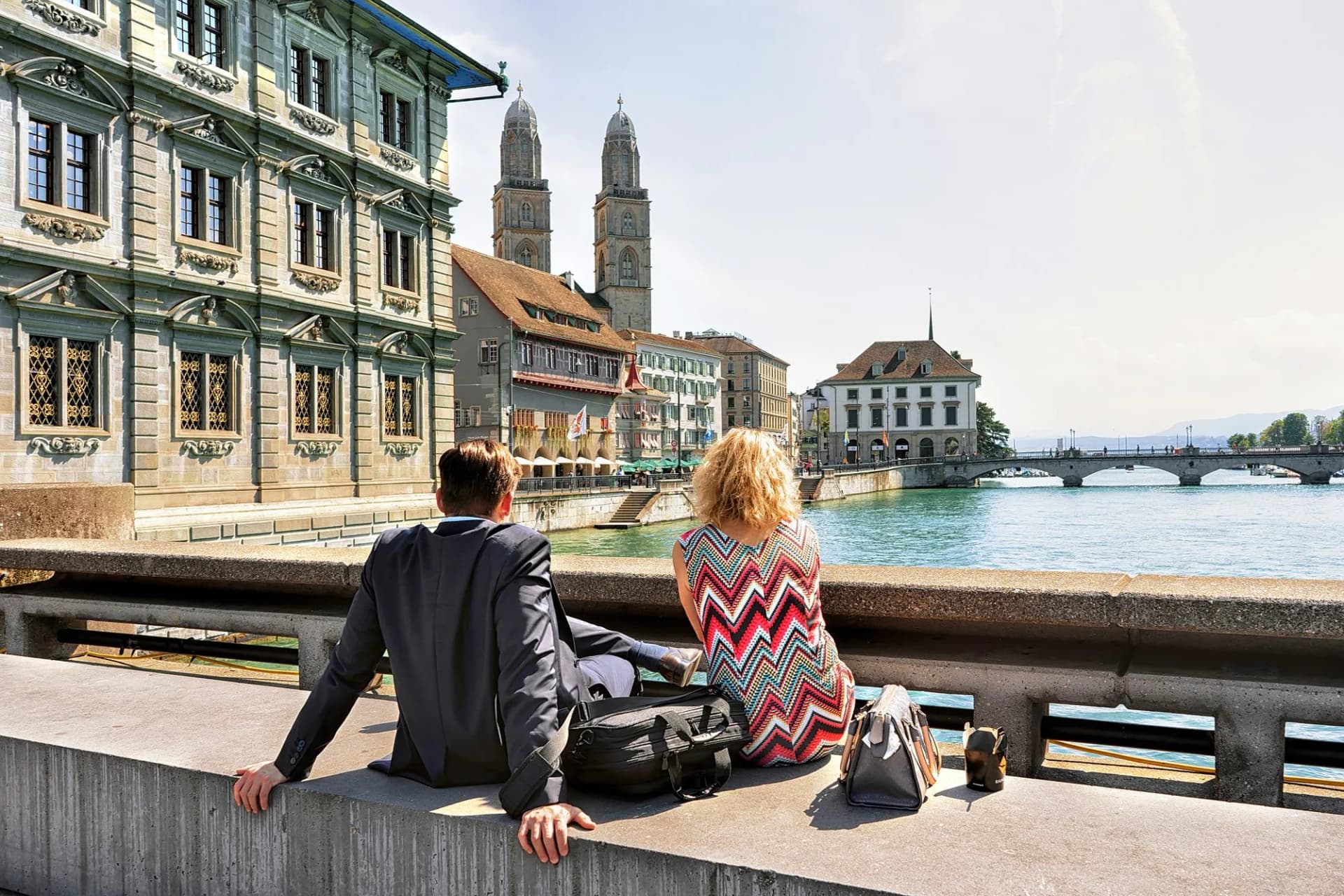 Couple sitting by Limmat River looking at Grossmünster church in Zurich, Switzerland.