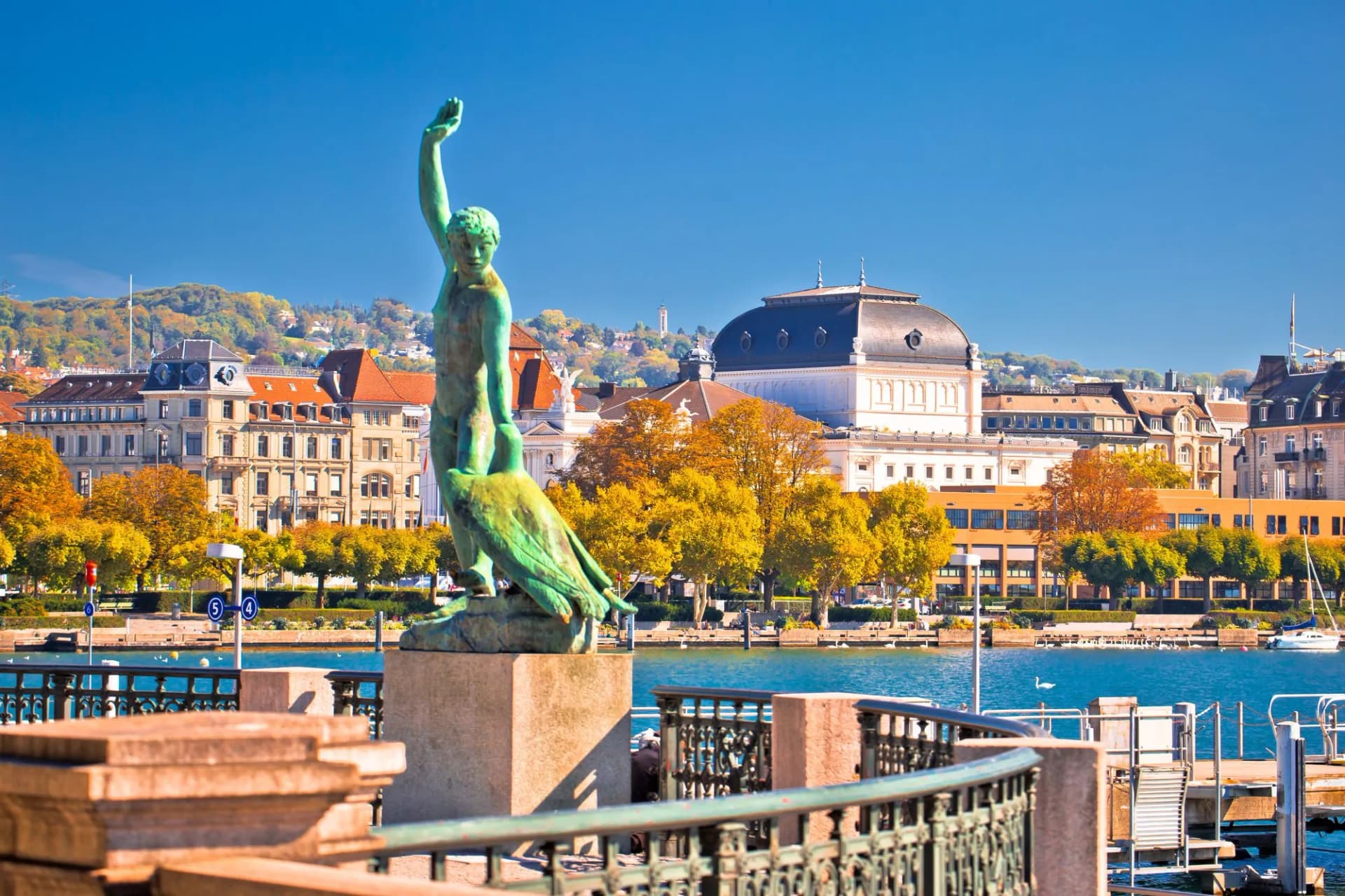 Bronze statue by Zurich Lake with autumn foliage and city buildings.