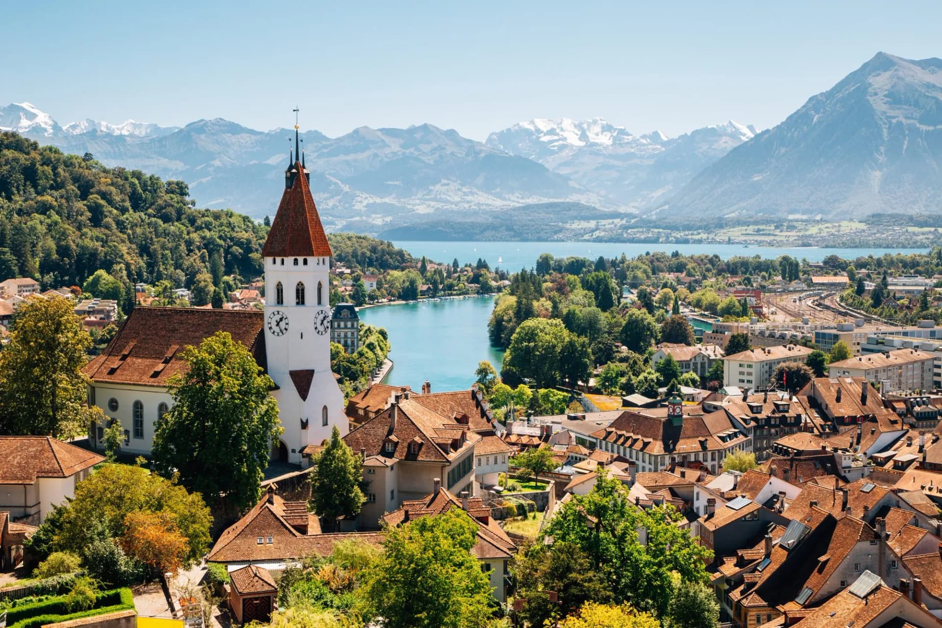 Thun cityscape with white church tower, Lake Thun, and snow-capped Alps mountains in Switzerland.