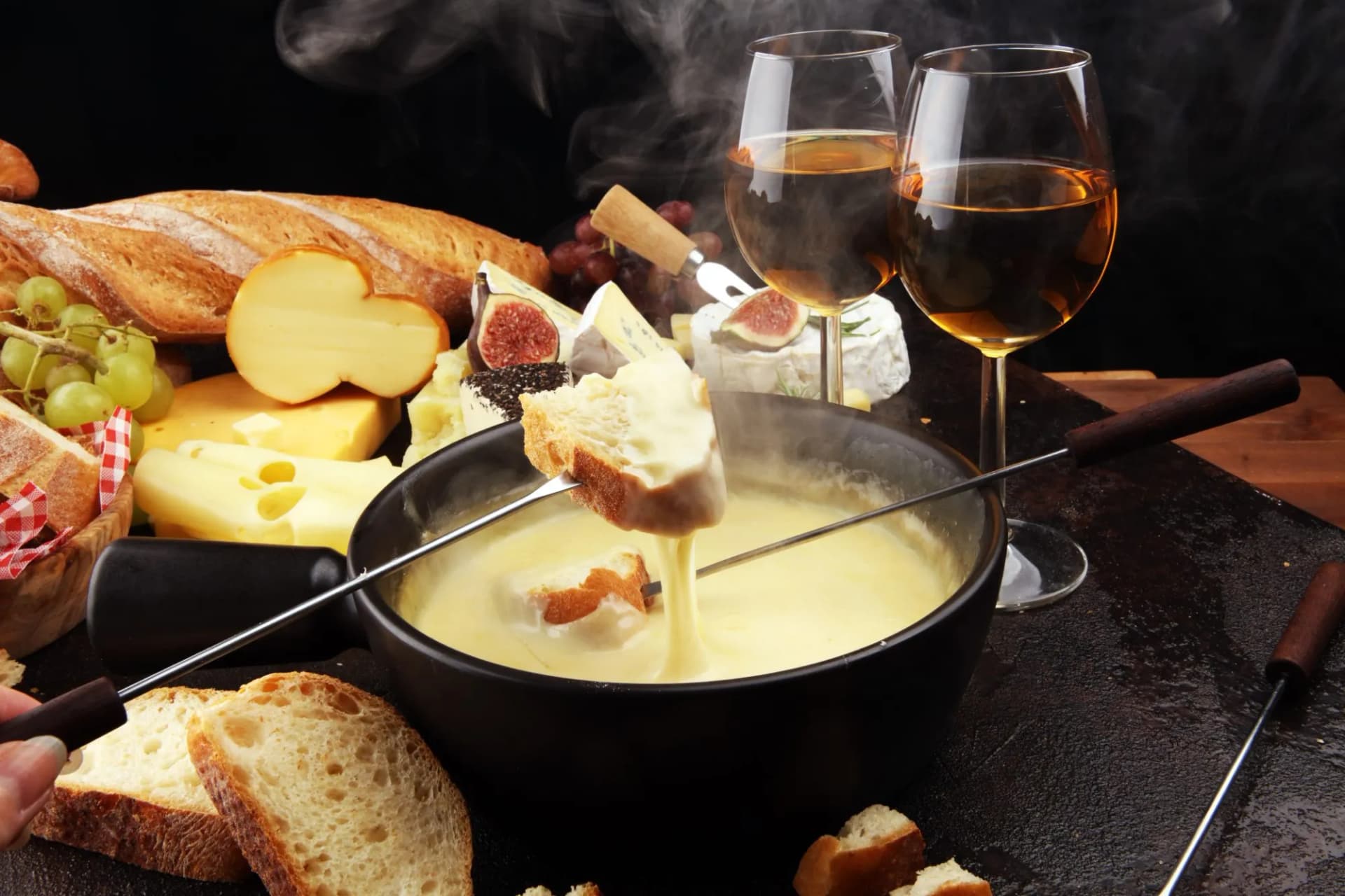 Cheese fondue dinner with bread, assorted cheeses, figs, grapes, and white wine on a dark surface.