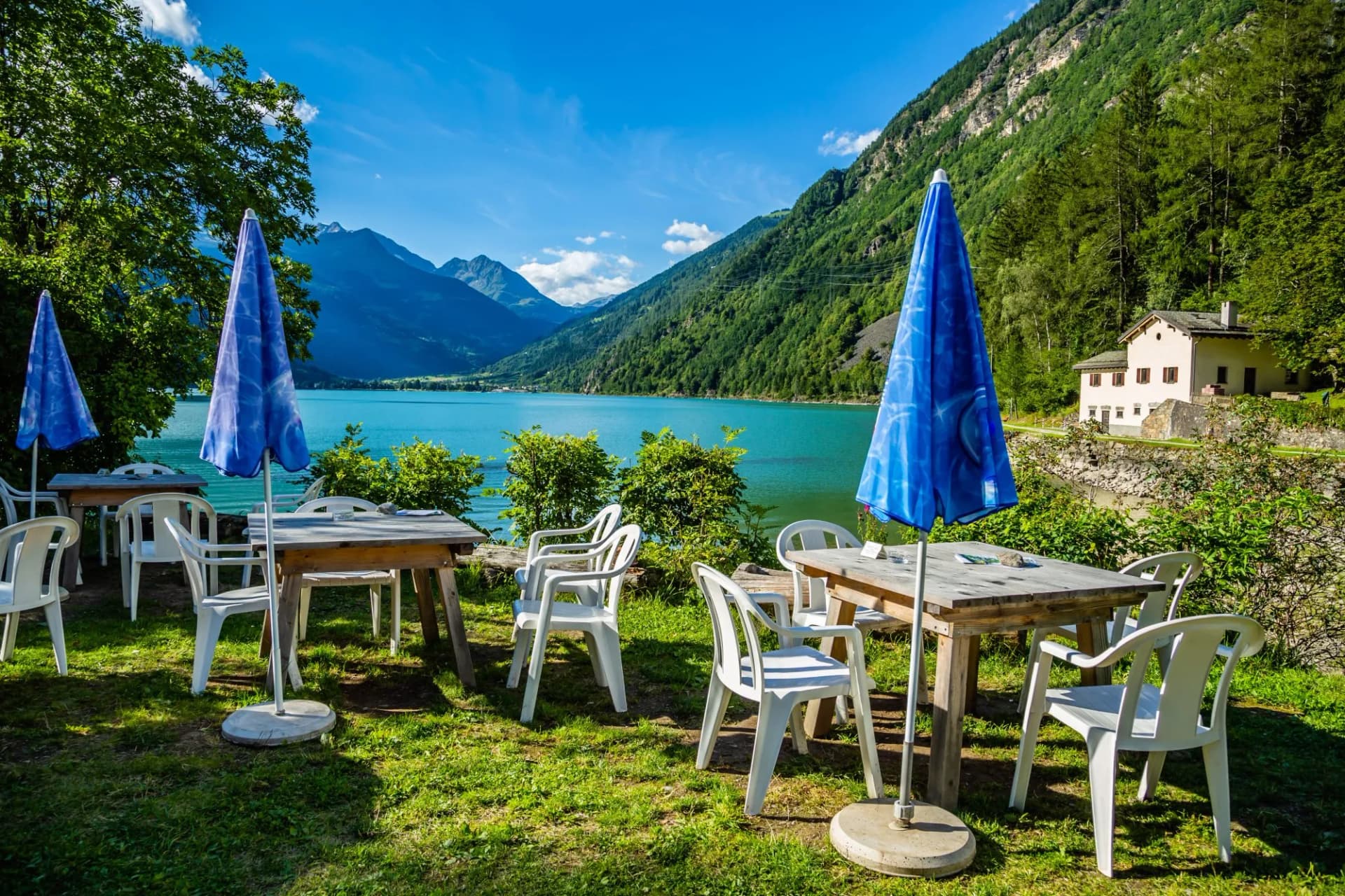Outdoor dining area with blue umbrellas overlooking Poschiavo Lake and green mountains.