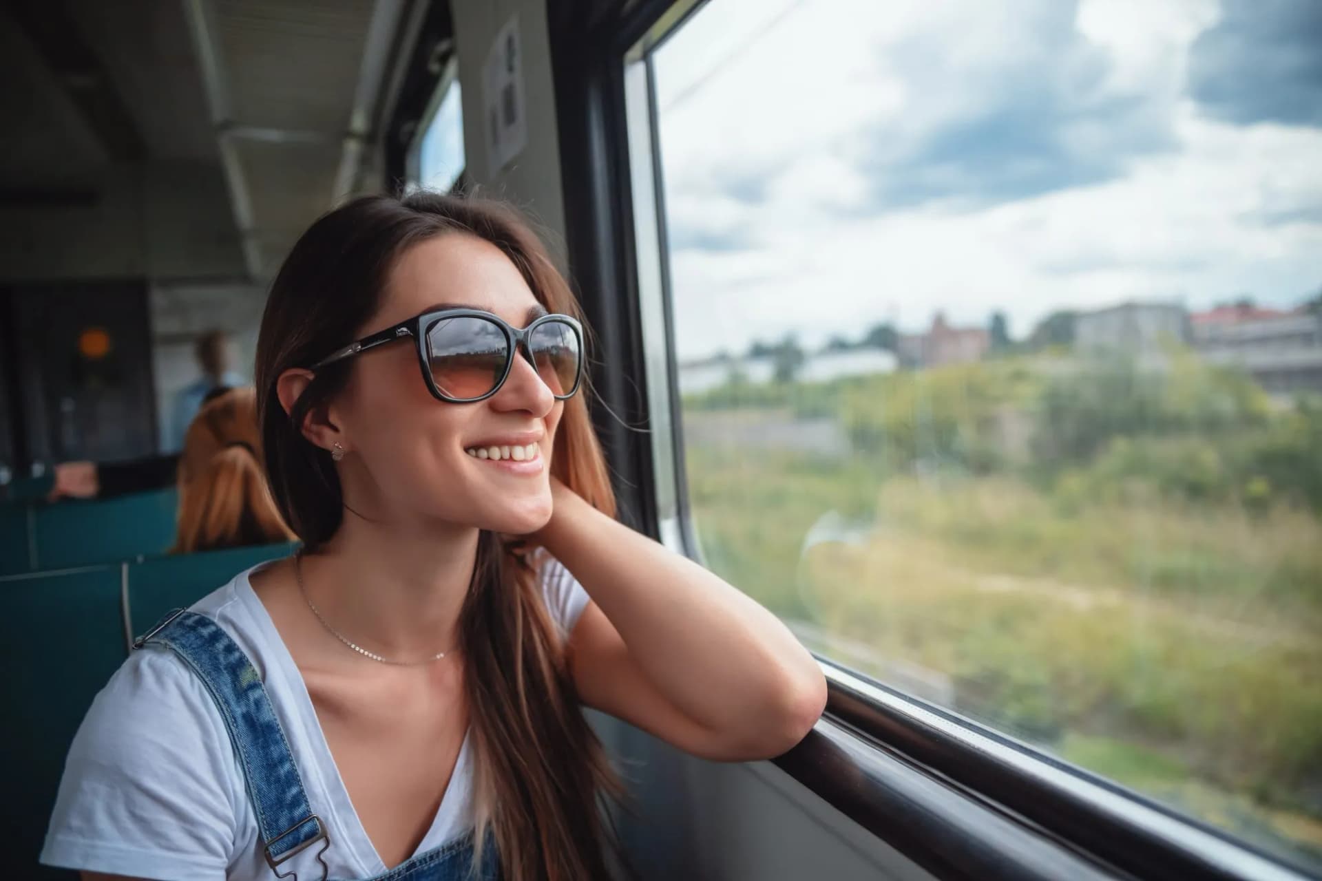 Young woman in sunglasses smiling while looking out the window during a train journey.