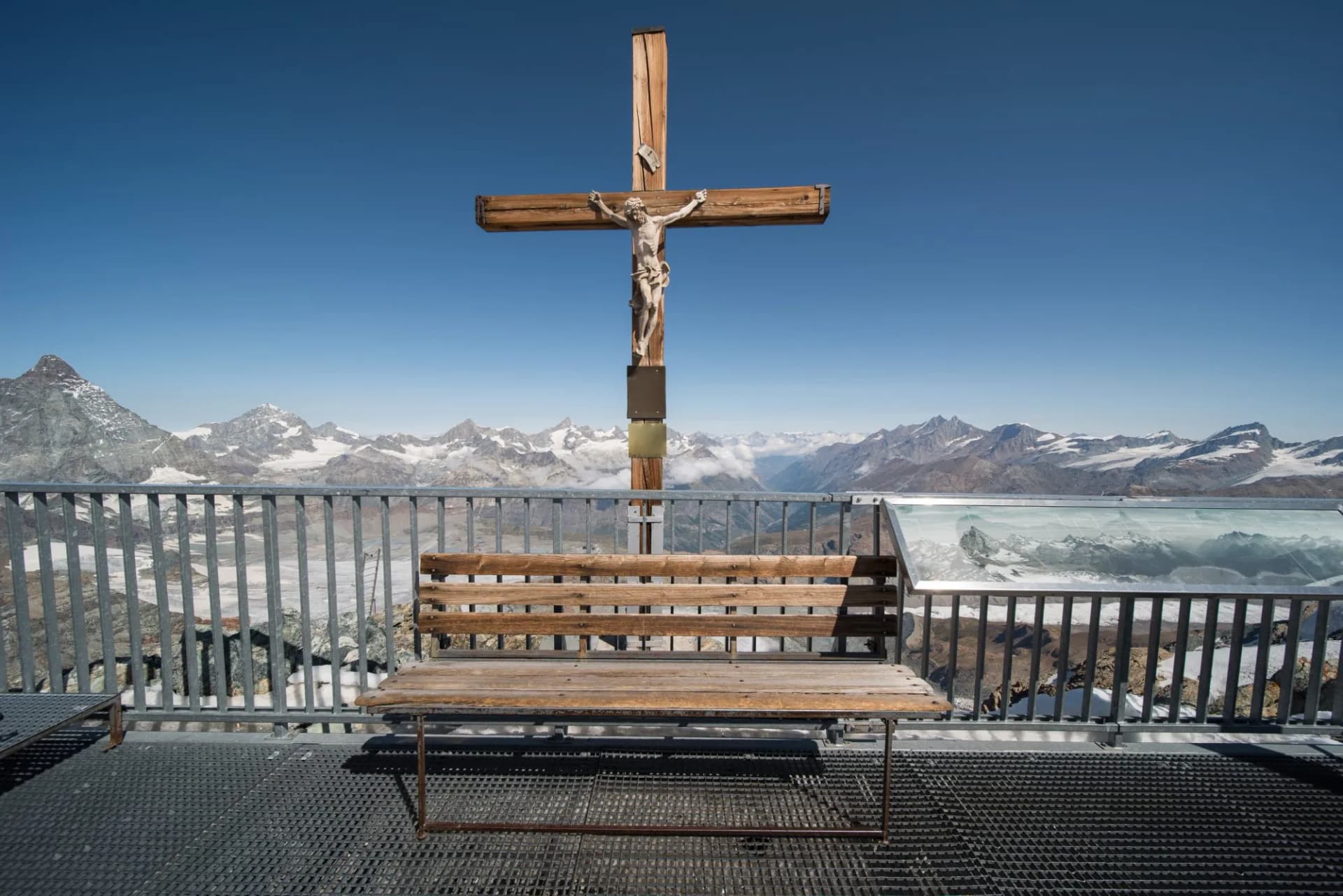 Summit cross and wooden bench on viewpoint overlooking snowy mountains near Zermatt, Switzerland.