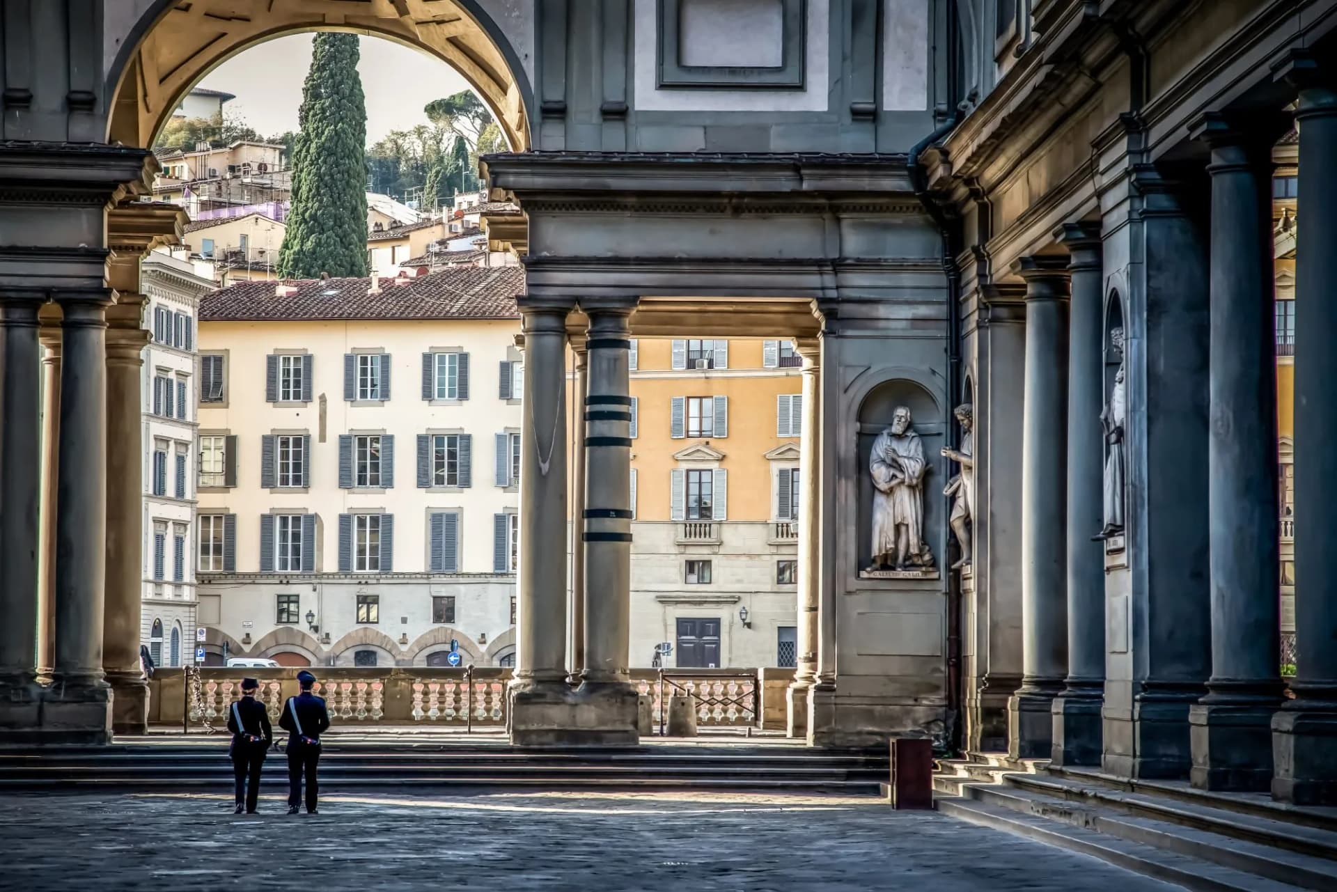 Uffizi Gallery colonnade with statues, two guards, and buildings visible in Piazza degli Uffizi, Florence.