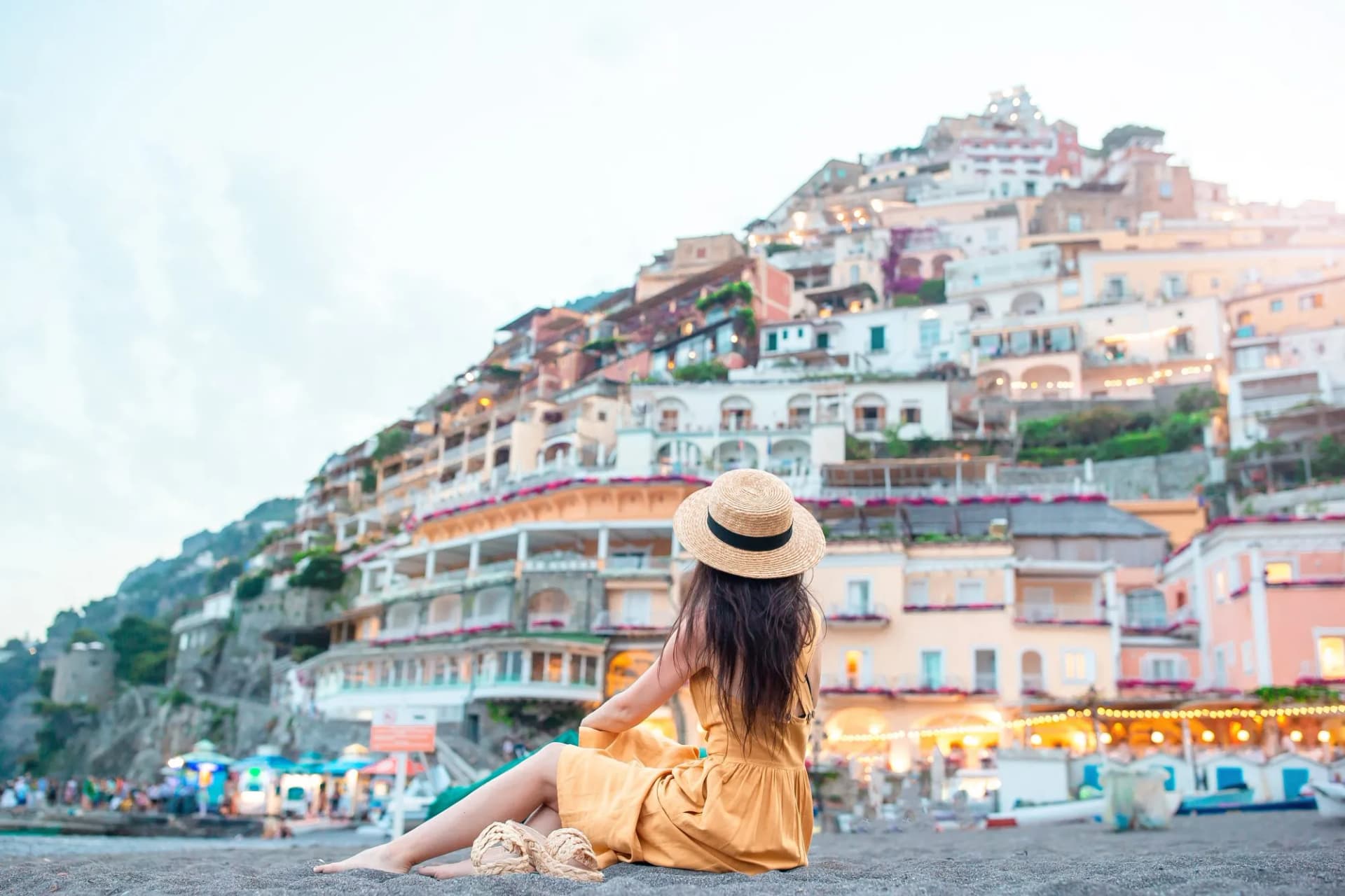 Woman in straw hat on beach facing colorful cliffside village of Positano, Amalfi Coast.