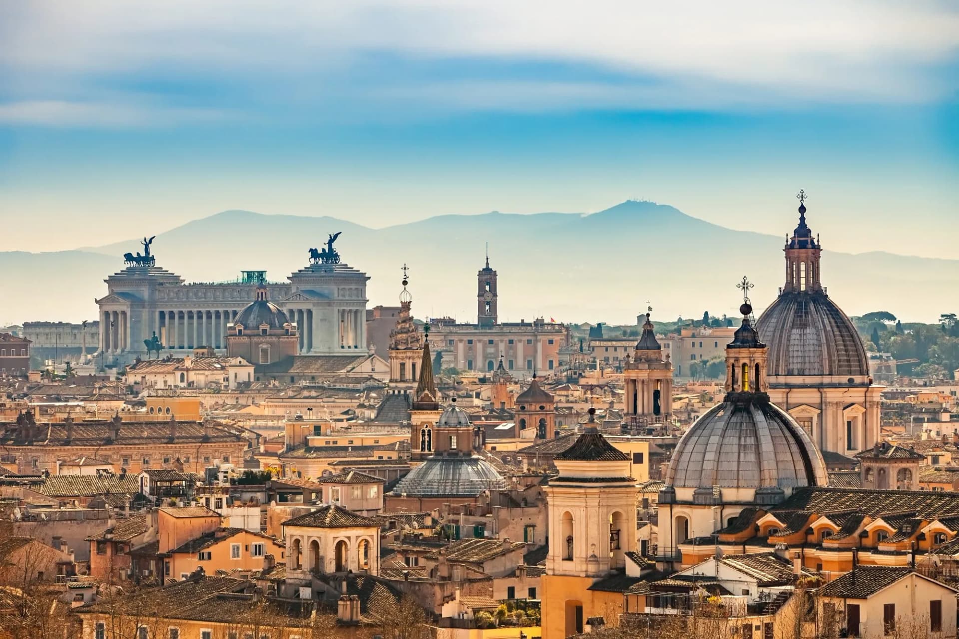 Rome skyline with domes and Vittoriano monument against hazy mountains and blue sky.
